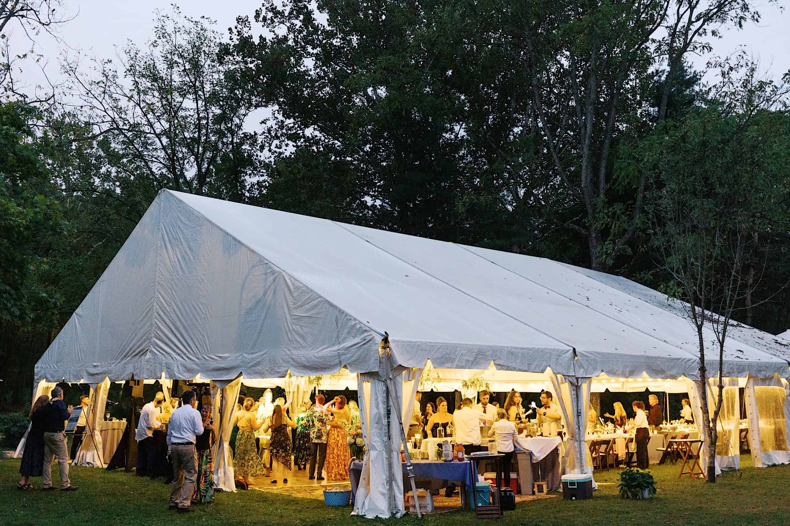A large white event tent is set up outdoors in a grassy area at dusk, where guests are gathered inside for a beautiful wedding at Vincent Forge Mansion.
