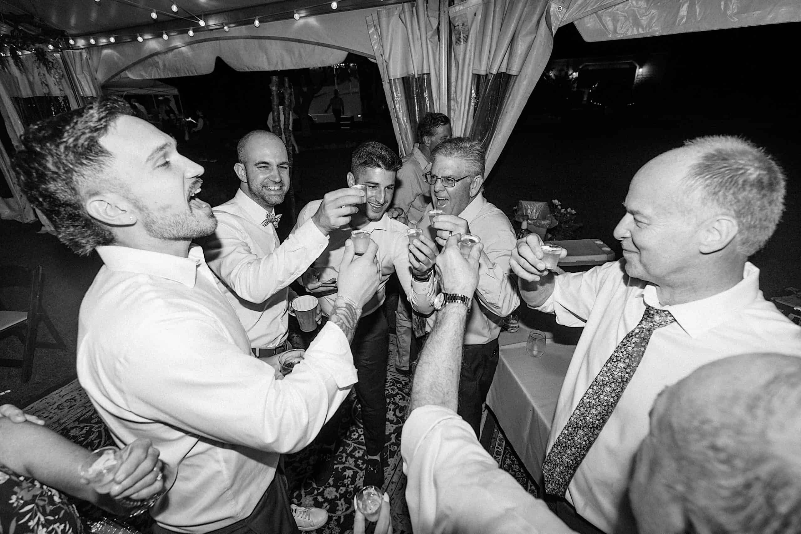 A group of men in dress shirts stand in a circle, raising small glasses in a toast under a tent at a nighttime event during a beautiful wedding at Vincent Forge Mansion.