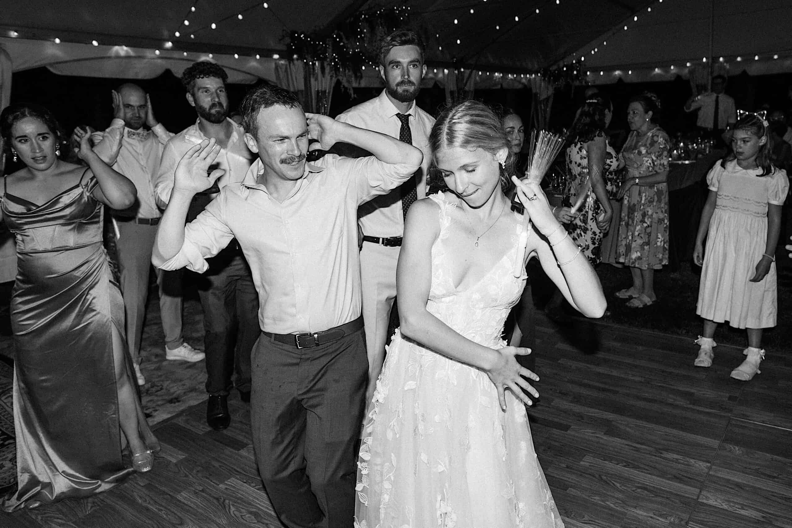 A group of people, including a bride in a white dress, dance energetically at a beautiful wedding at Vincent Forge Mansion under glowing string lights.