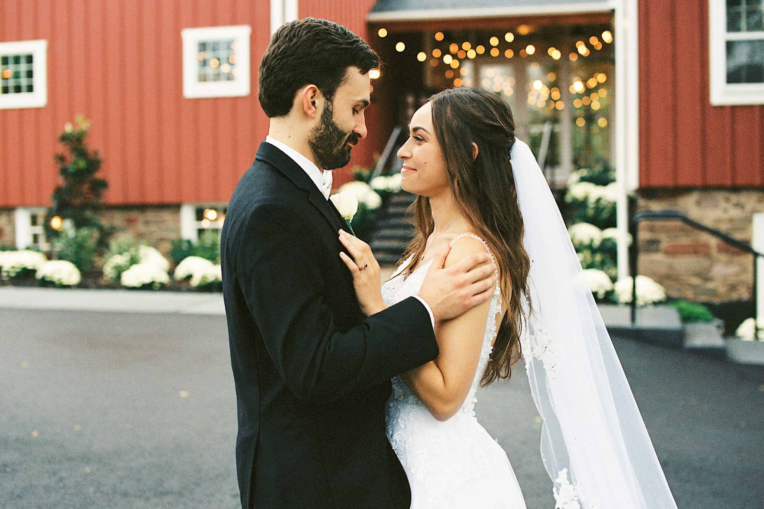 A bride and groom in formal attire stand facing each other outside a red barn decorated with string lights at a beautiful Lochwood Manor wedding.