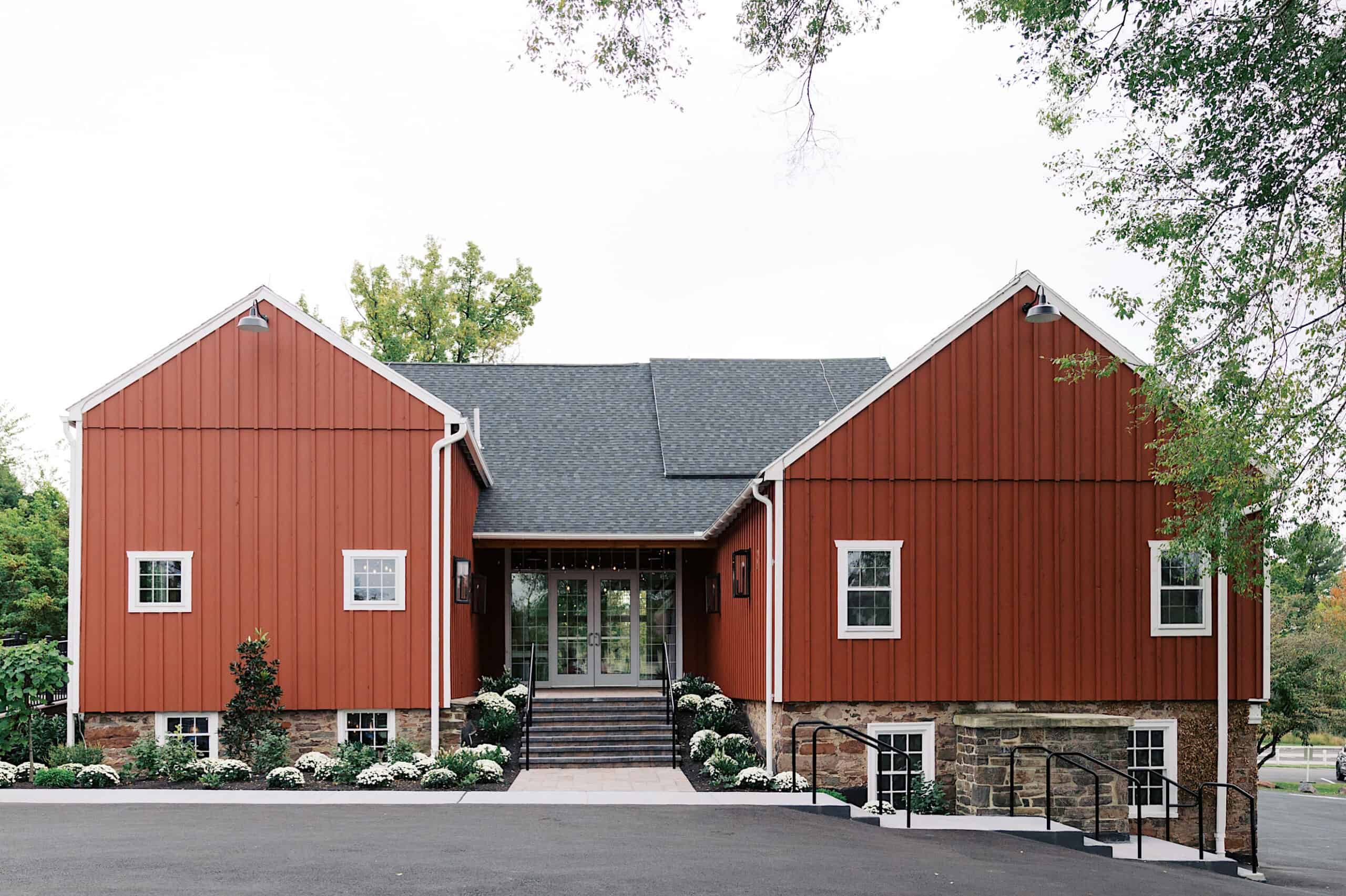 A modern red barn-style building with two wings, stone foundation, and central glass entrance, surrounded by greenery and white flowers—perfect for a beautiful Lochwood Manor wedding.
