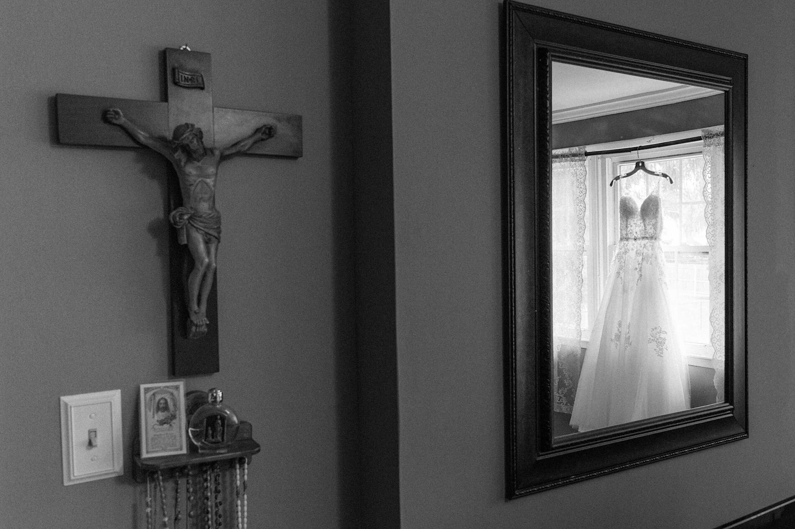 A crucifix hangs on the wall beside a mirror reflecting a wedding dress on a hanger in front of a window, capturing the anticipation of a beautiful Lochwood Manor wedding.