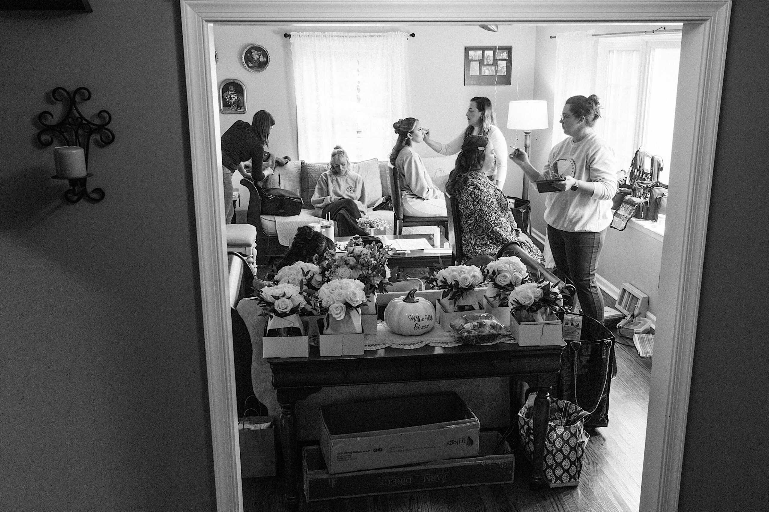 A group of women prepares in a living room for a beautiful Lochwood Manor wedding, with some getting their hair and makeup done. Gift bags and floral arrangements are arranged on a table in the foreground.