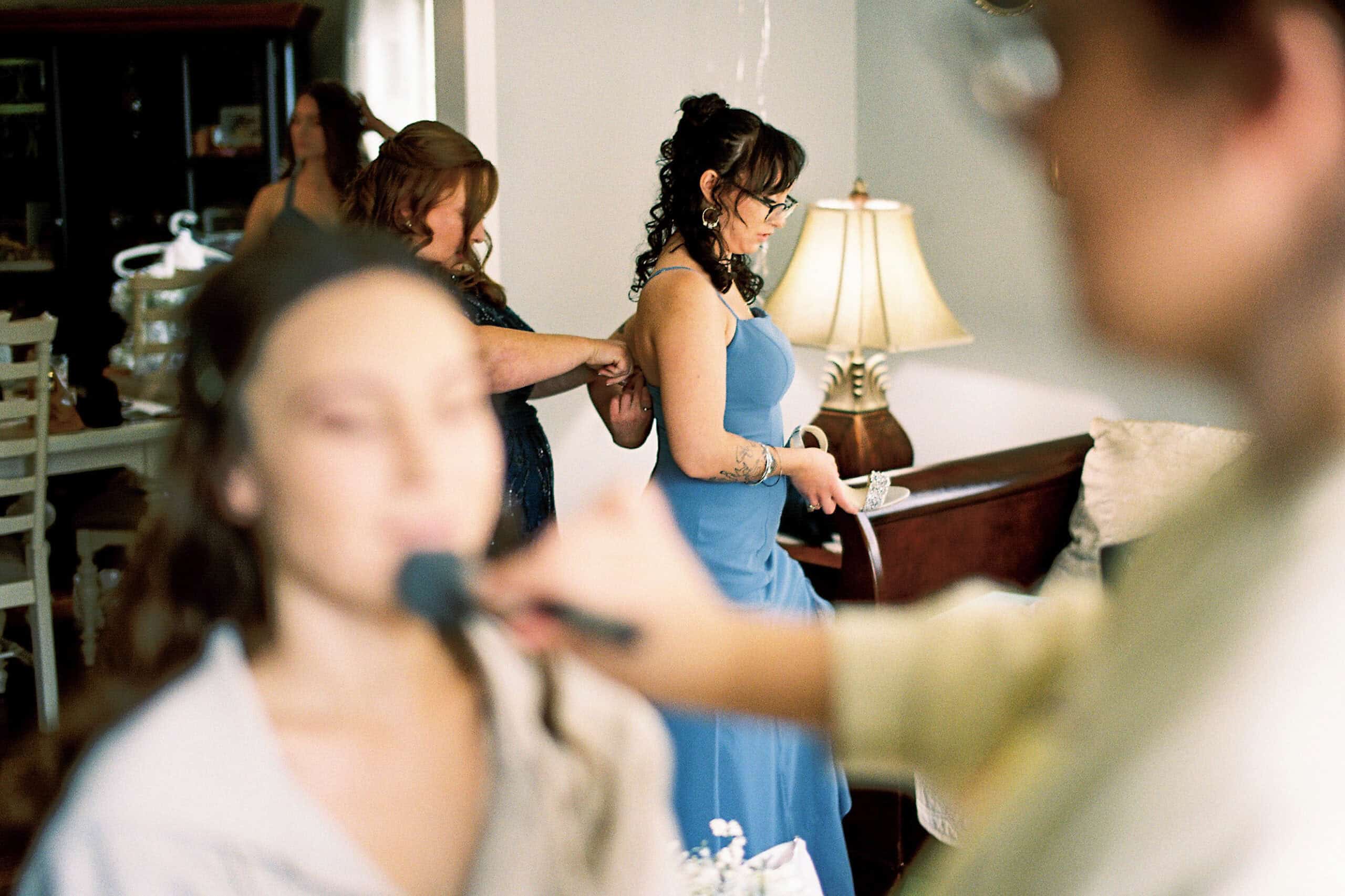 A woman in a blue dress stands still as another adjusts her dress at a beautiful Lochwood Manor wedding, while a blurred figure in the foreground has makeup applied. A lamp and furniture are visible in the background.