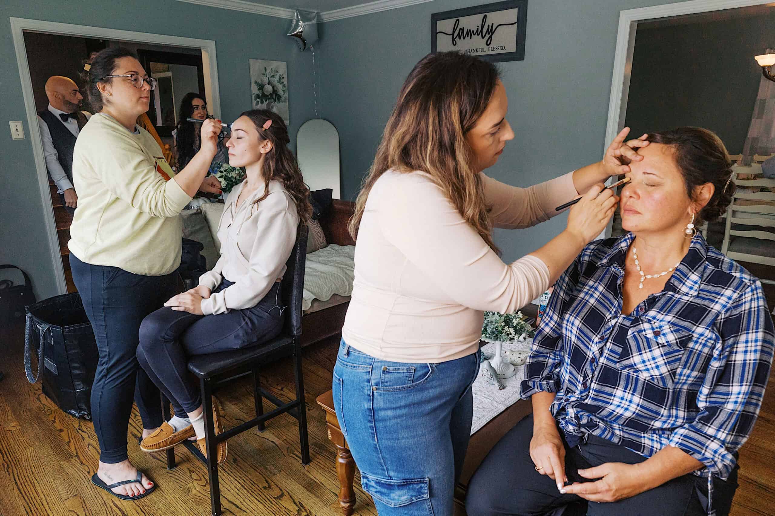Two makeup artists apply makeup to two women seated in a living room, preparing for a beautiful Lochwood Manor wedding, while two other people stand in the background.