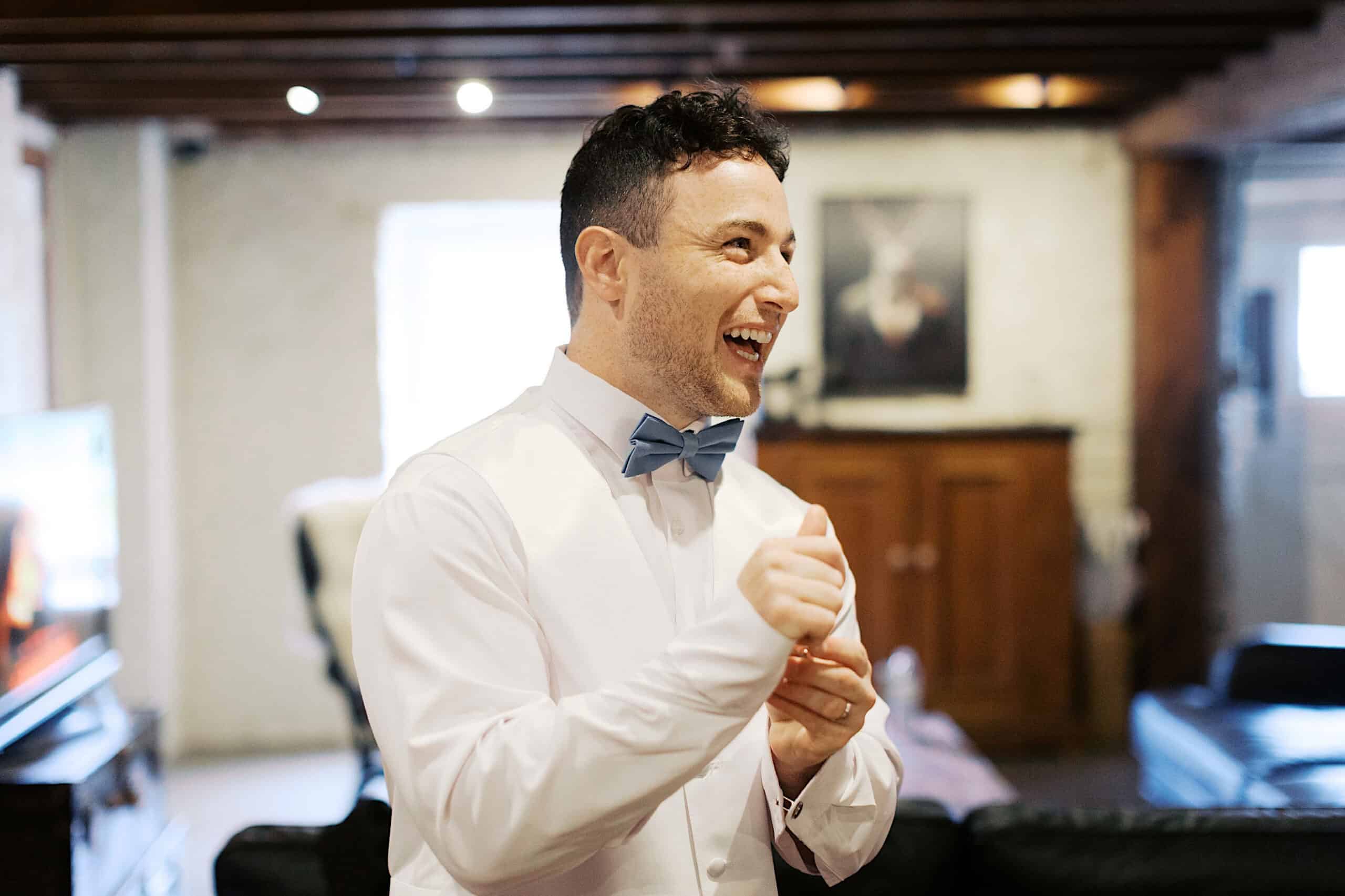 A man in a white shirt and blue bow tie smiles and laughs in a living room with wooden ceiling beams at a beautiful Lochwood Manor wedding.