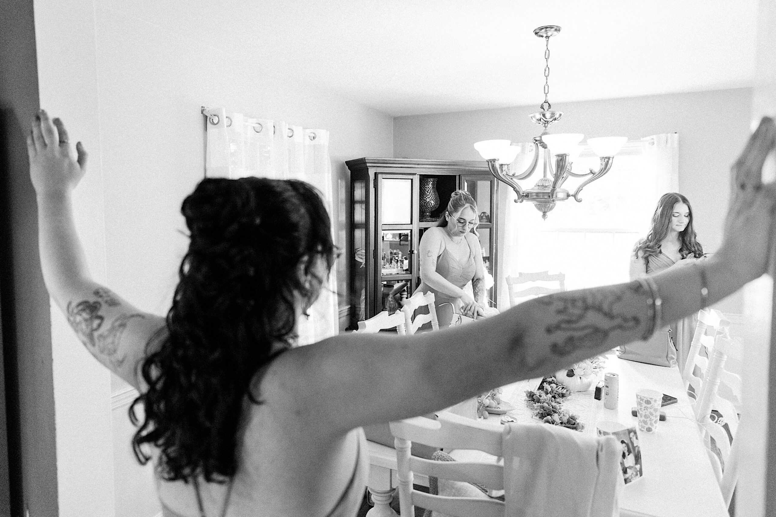 A woman with tattoos stands with arms outstretched in the foreground, while two other women prepare at a dining table in a well-lit room before a beautiful Lochwood Manor wedding.