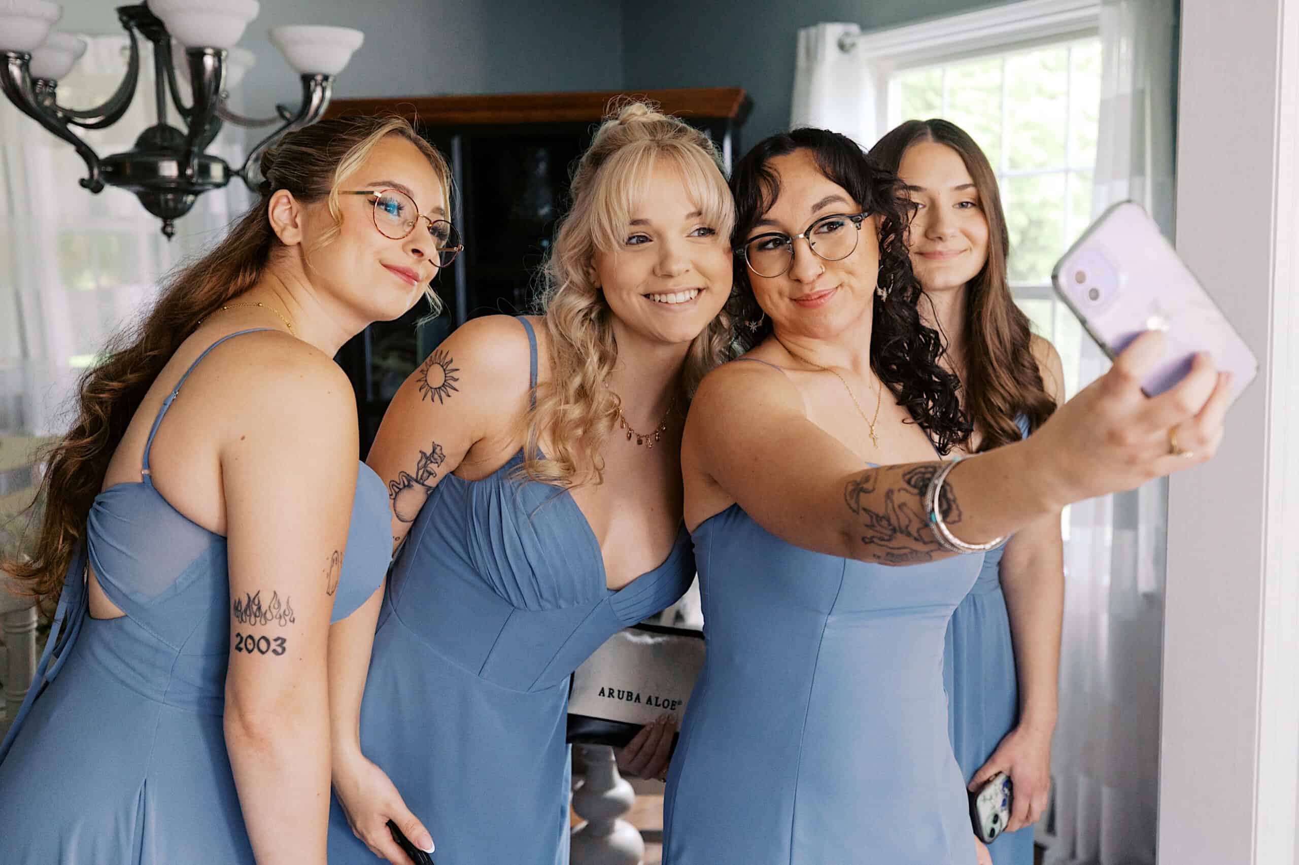 Four women in matching blue dresses stand close together and smile as one of them takes a selfie with a smartphone at a beautiful Lochwood Manor wedding in a well-lit room.