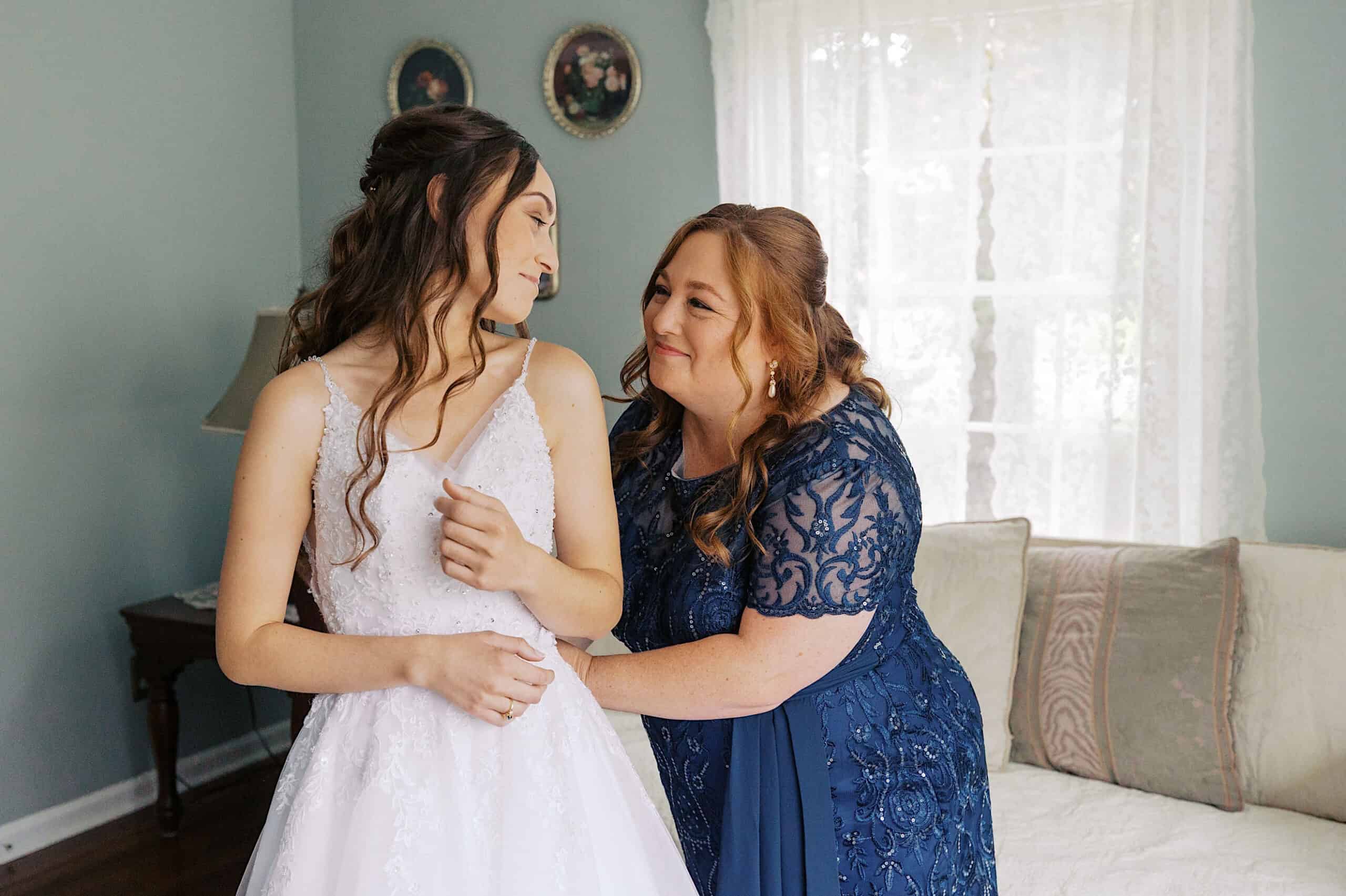 A woman in a blue dress helps a bride adjust her gown in a softly lit room with pale blue walls and framed pictures at a beautiful Lochwood Manor wedding.