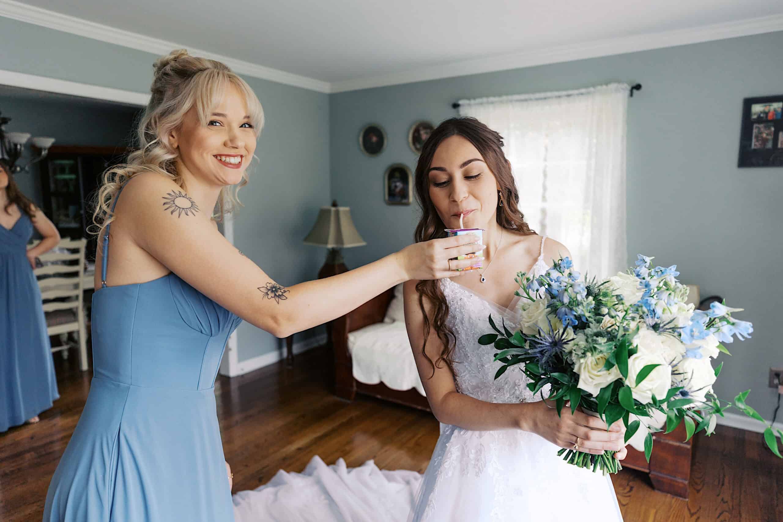 At a beautiful Lochwood Manor wedding, a bridesmaid in a blue dress helps the bride, who is holding a bouquet, drink from a cup in a sunlit room.