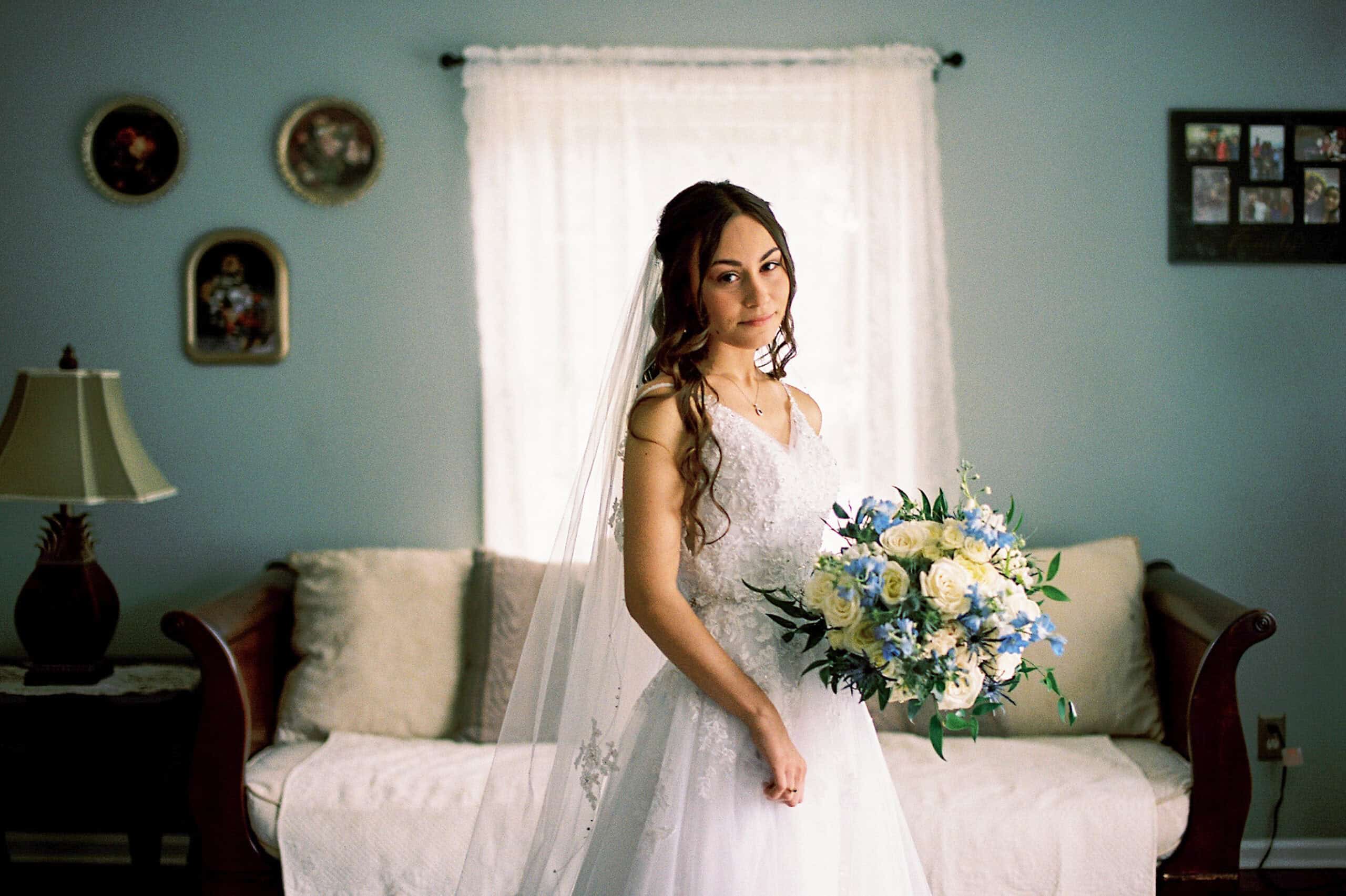 A bride in a white gown and veil stands indoors at a beautiful Lochwood Manor wedding, holding a bouquet of white and blue flowers, with a sofa and framed pictures in the background.