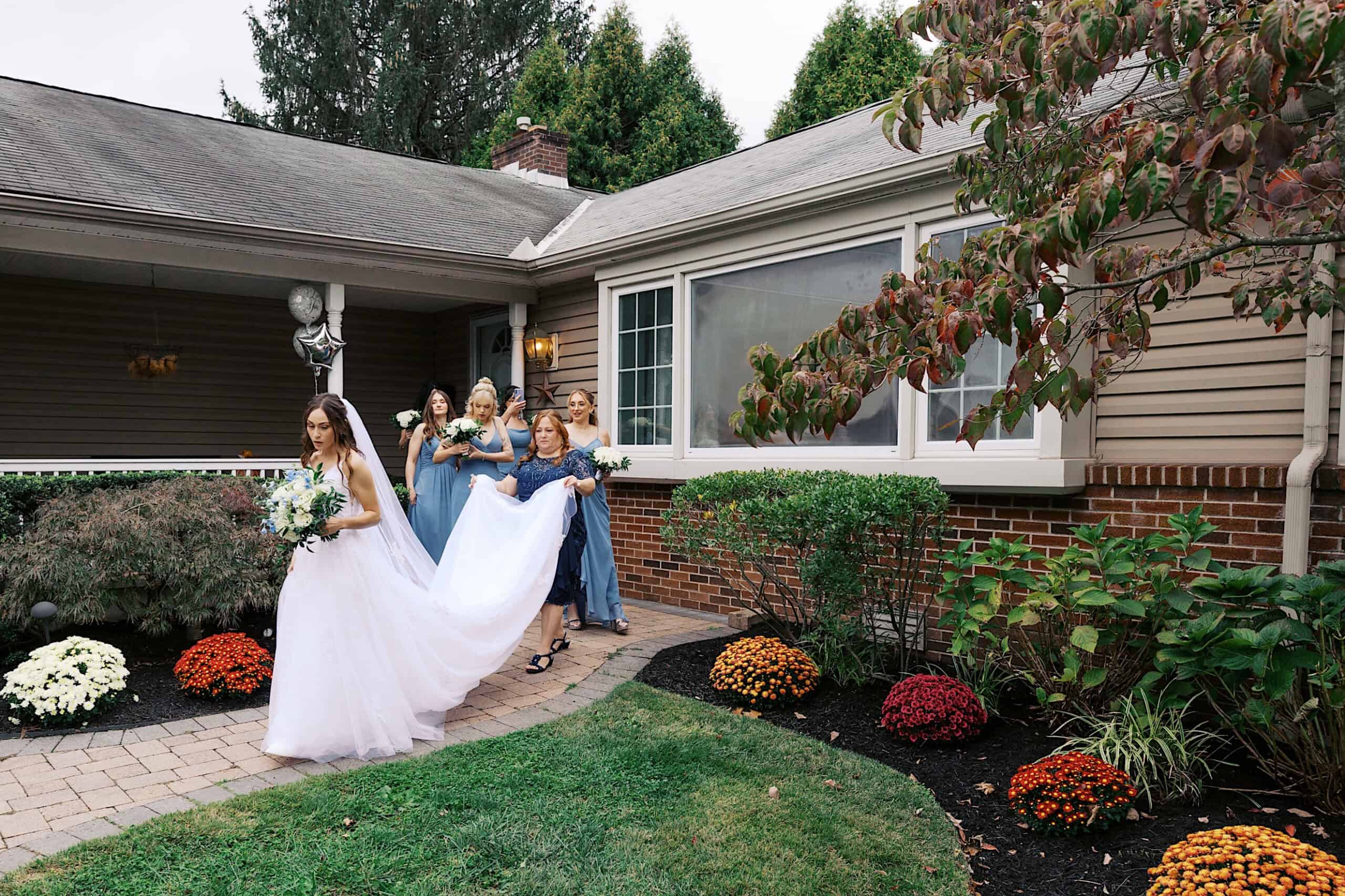 A bride in a white dress walks on a garden path outside Lochwood Manor, followed by attendants in blue dresses helping with her train at this beautiful wedding.
