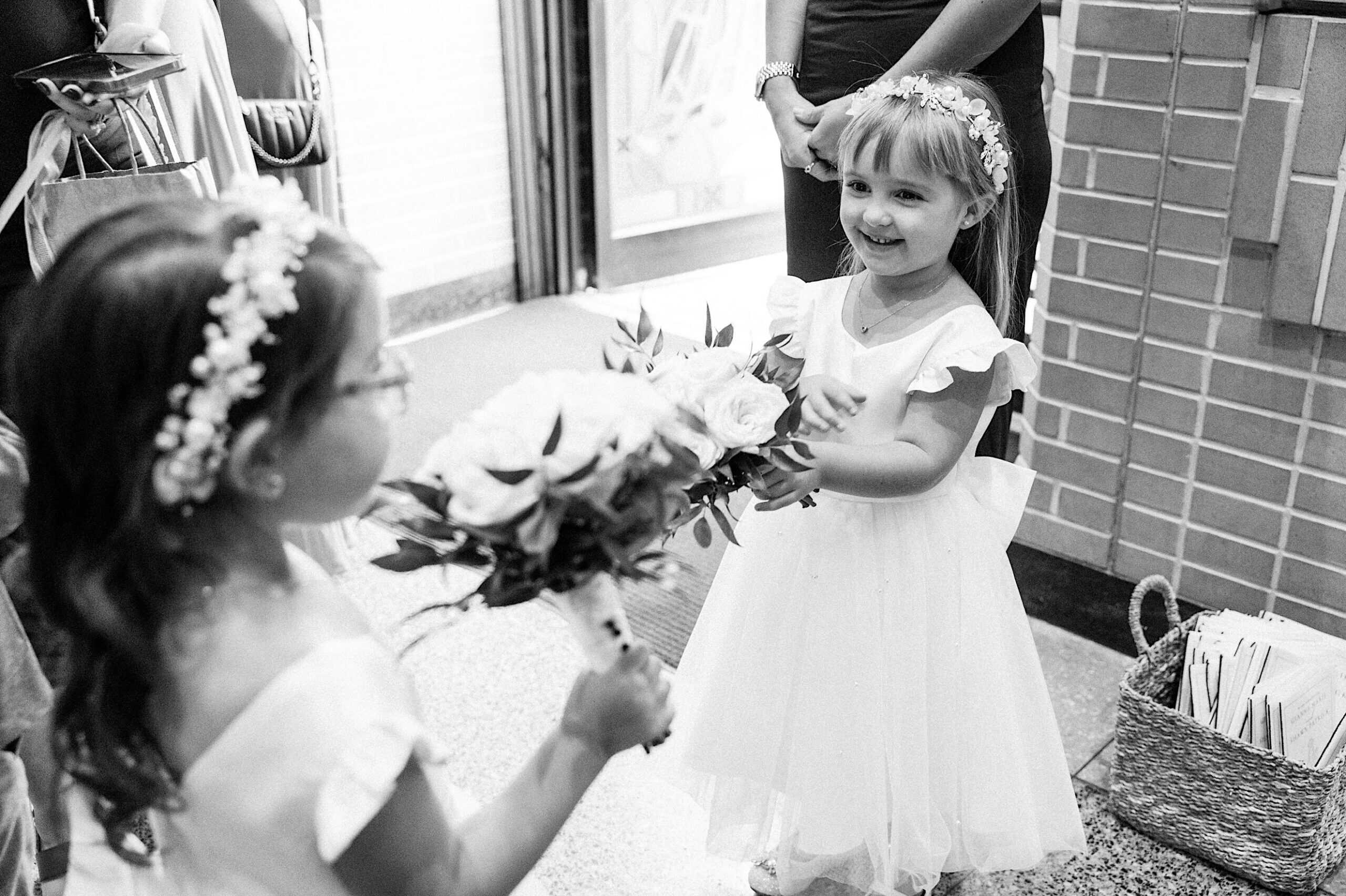 Two young girls in white dresses and floral headbands stand indoors at a beautiful Lochwood Manor wedding, one holding a bouquet of flowers while the other smiles.