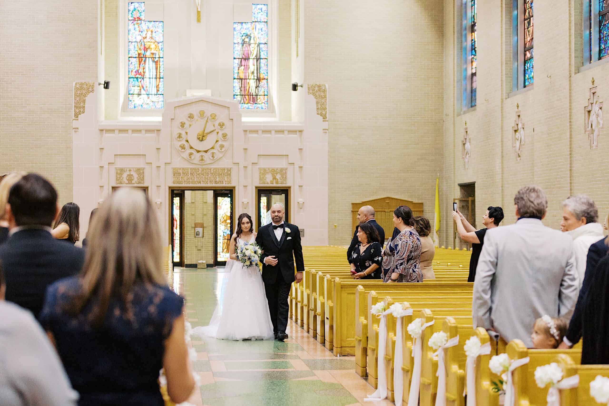 A bride in a white dress walks down the aisle with a man in a suit inside Lochwood Manor, as guests seated in pews watch and take photos at this beautiful wedding.