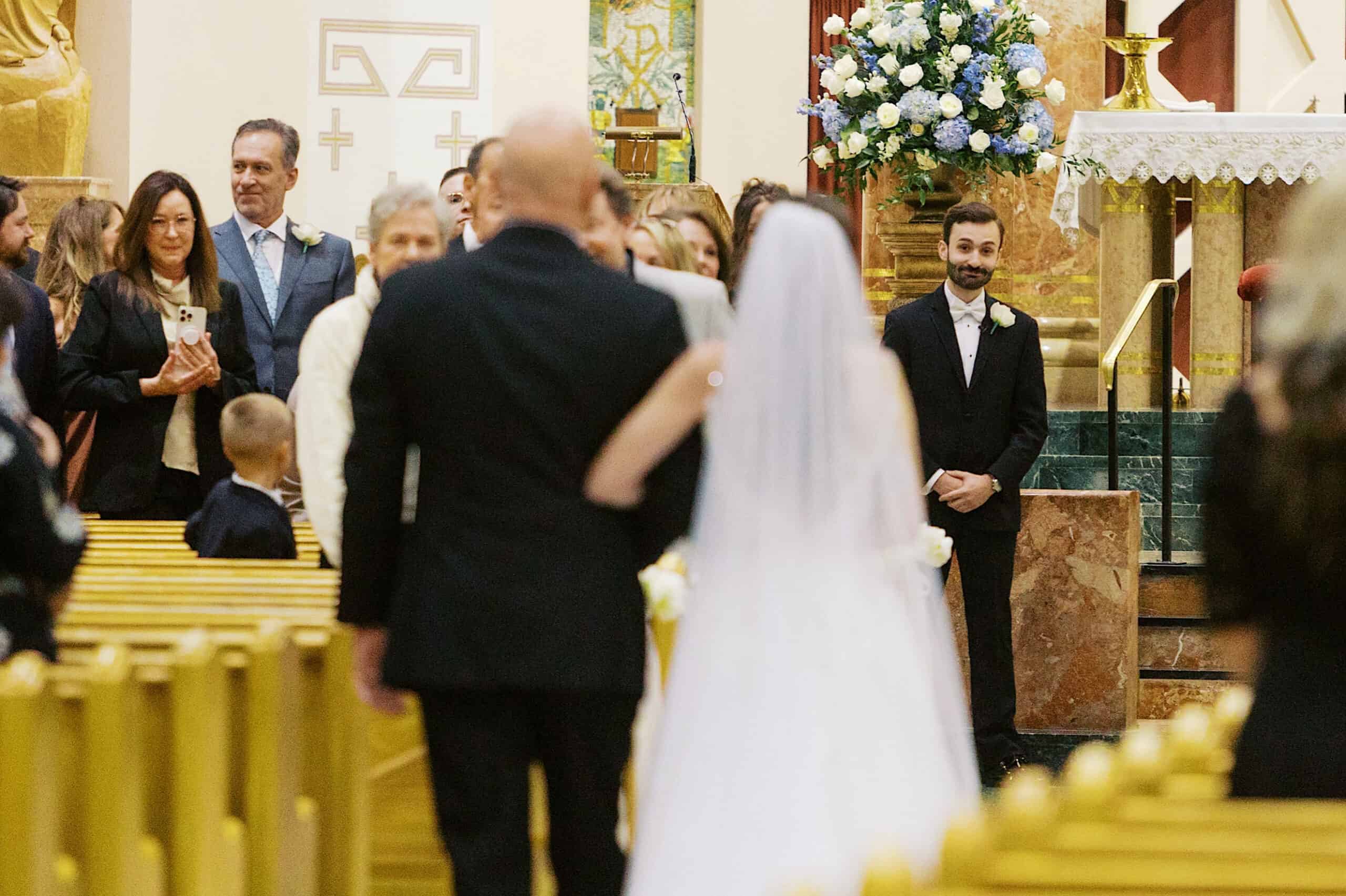 A bride in a white dress walks down the aisle with a man in a black suit toward a groom waiting at the altar during a beautiful Lochwood Manor wedding, surrounded by seated and standing guests in the church.