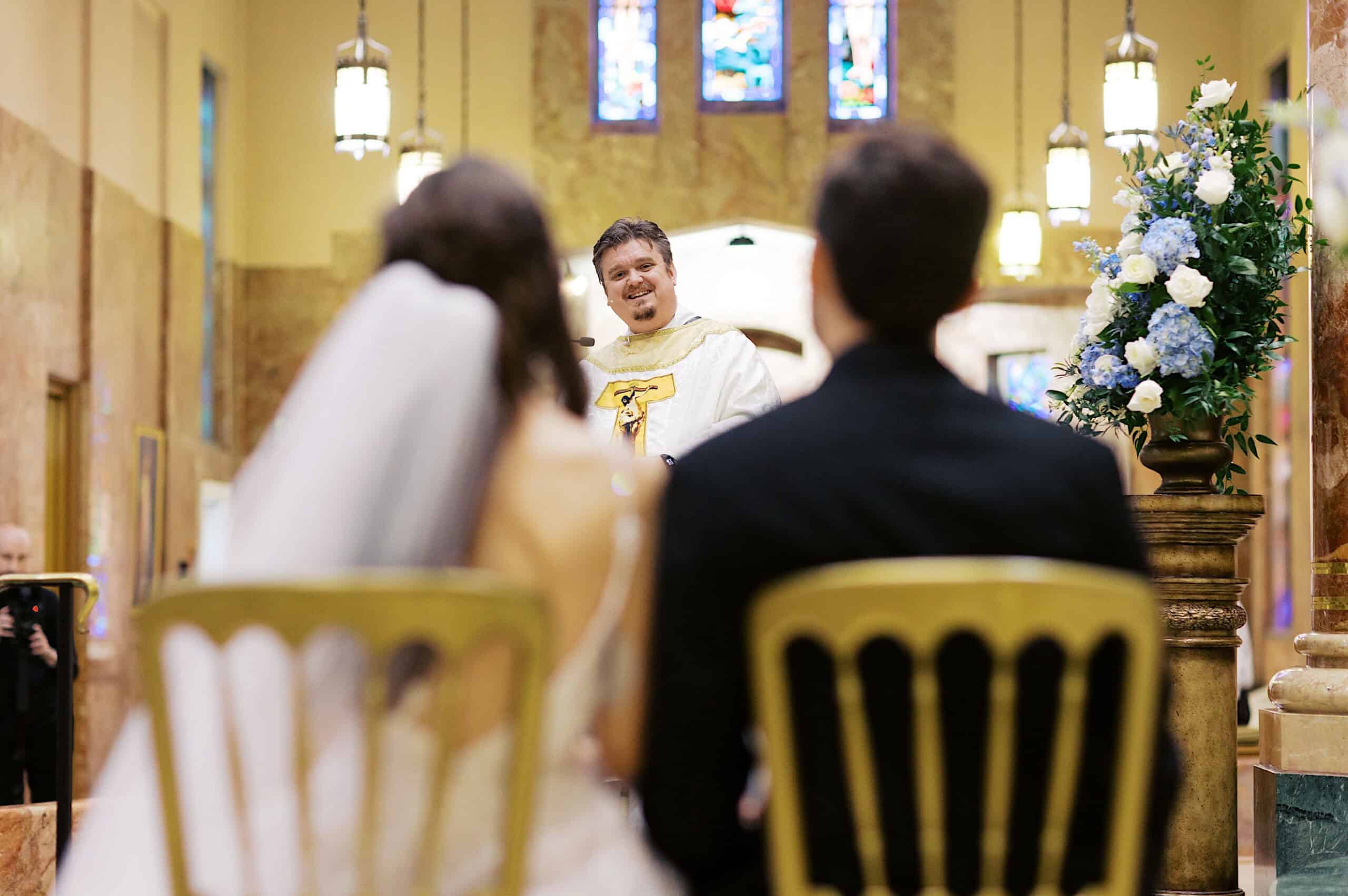 A priest stands at the altar speaking to a bride and groom seated in a church decorated with flowers and stained glass windows, creating a beautiful Lochwood Manor wedding scene.