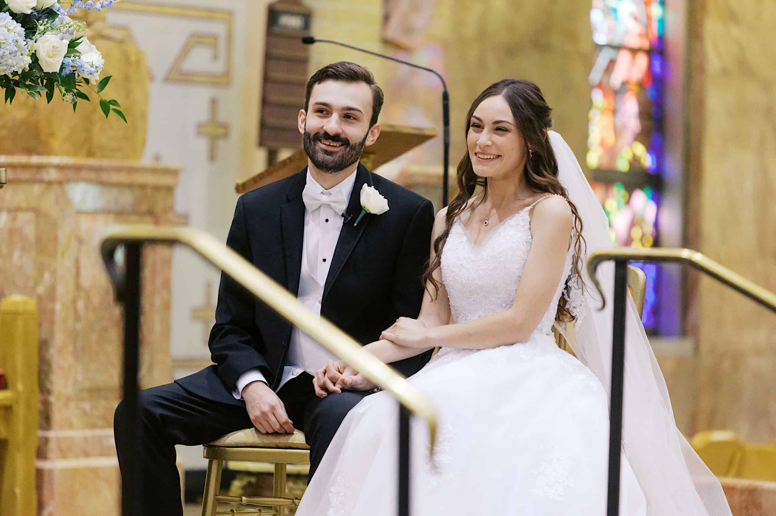 A bride and groom sit together in formal attire, holding hands and smiling inside a church with colorful stained glass in the background at their beautiful Lochwood Manor wedding.