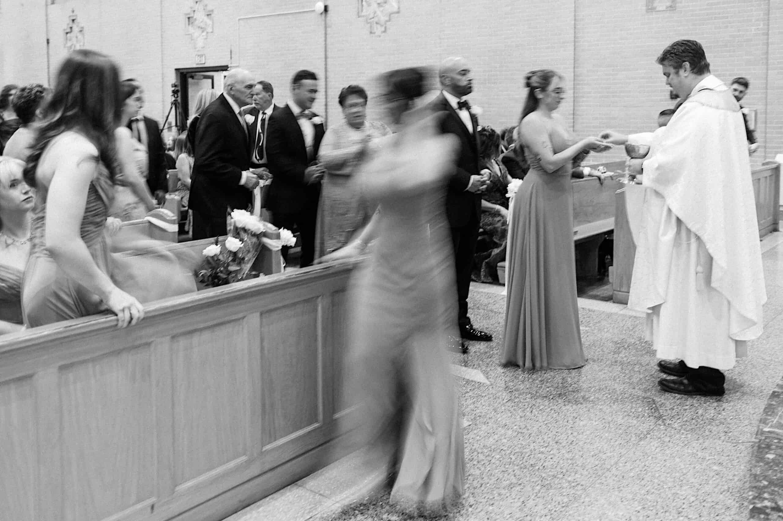 Black-and-white photo of a church interior at a beautiful Lochwood Manor wedding, showing a priest giving communion to people in formal attire; some figures are blurred in motion.