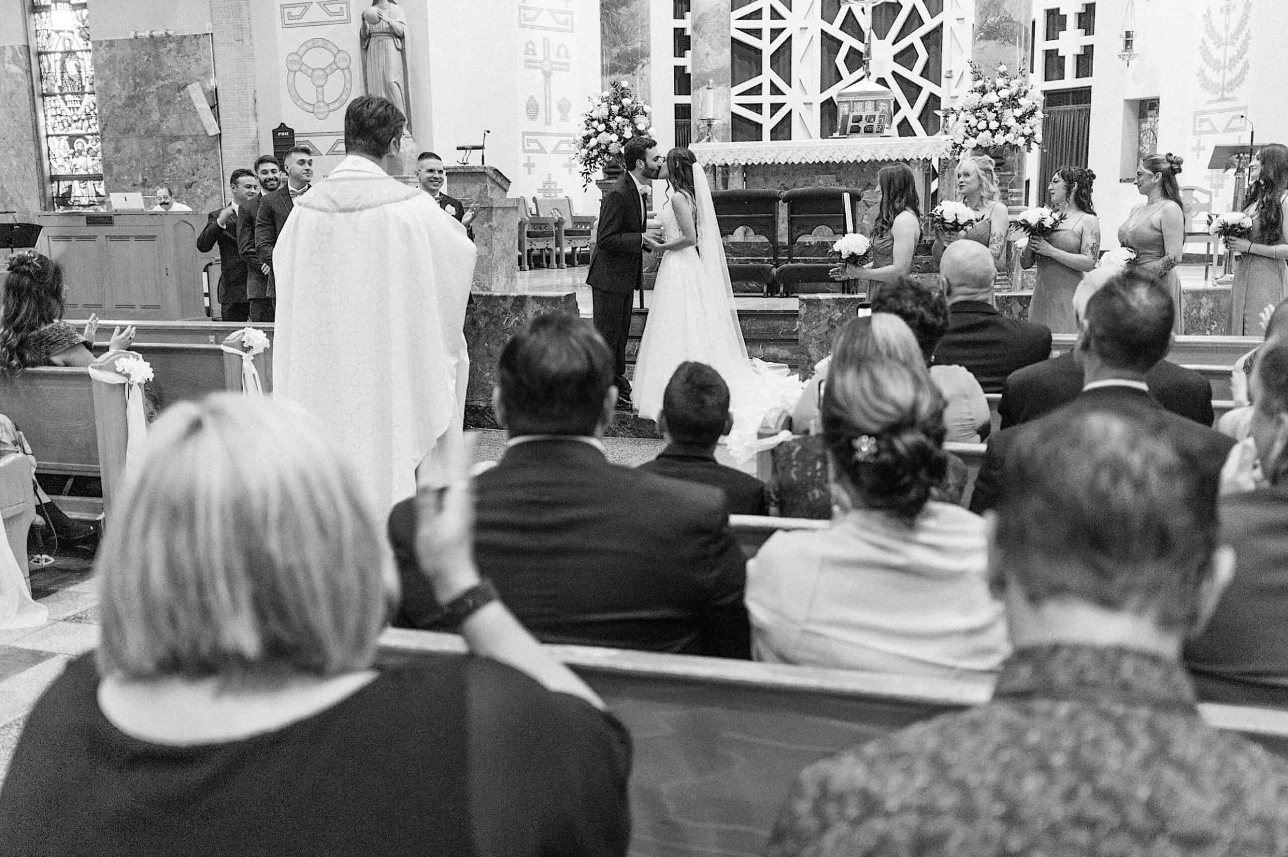 A bride and groom stand at the altar during a beautiful Lochwood Manor wedding ceremony in a church, surrounded by the bridal party, guests seated in pews, and a priest officiating.