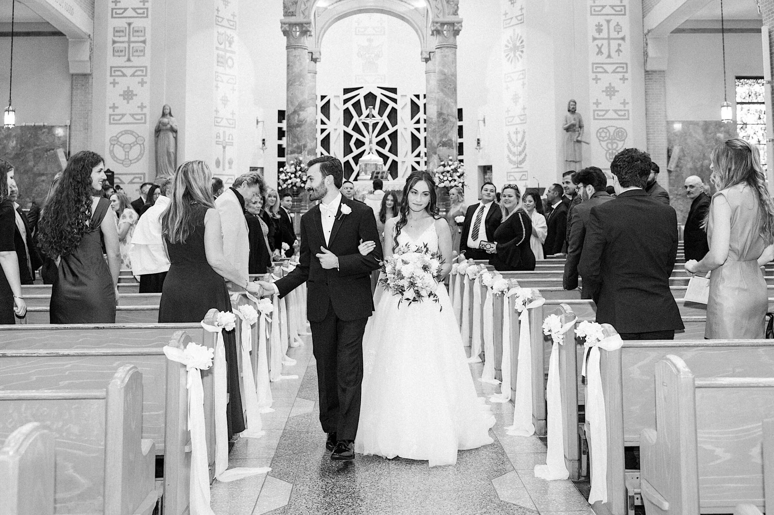 A bride and groom walk down the aisle together in a church during their beautiful Lochwood Manor wedding, surrounded by guests who are standing and watching.