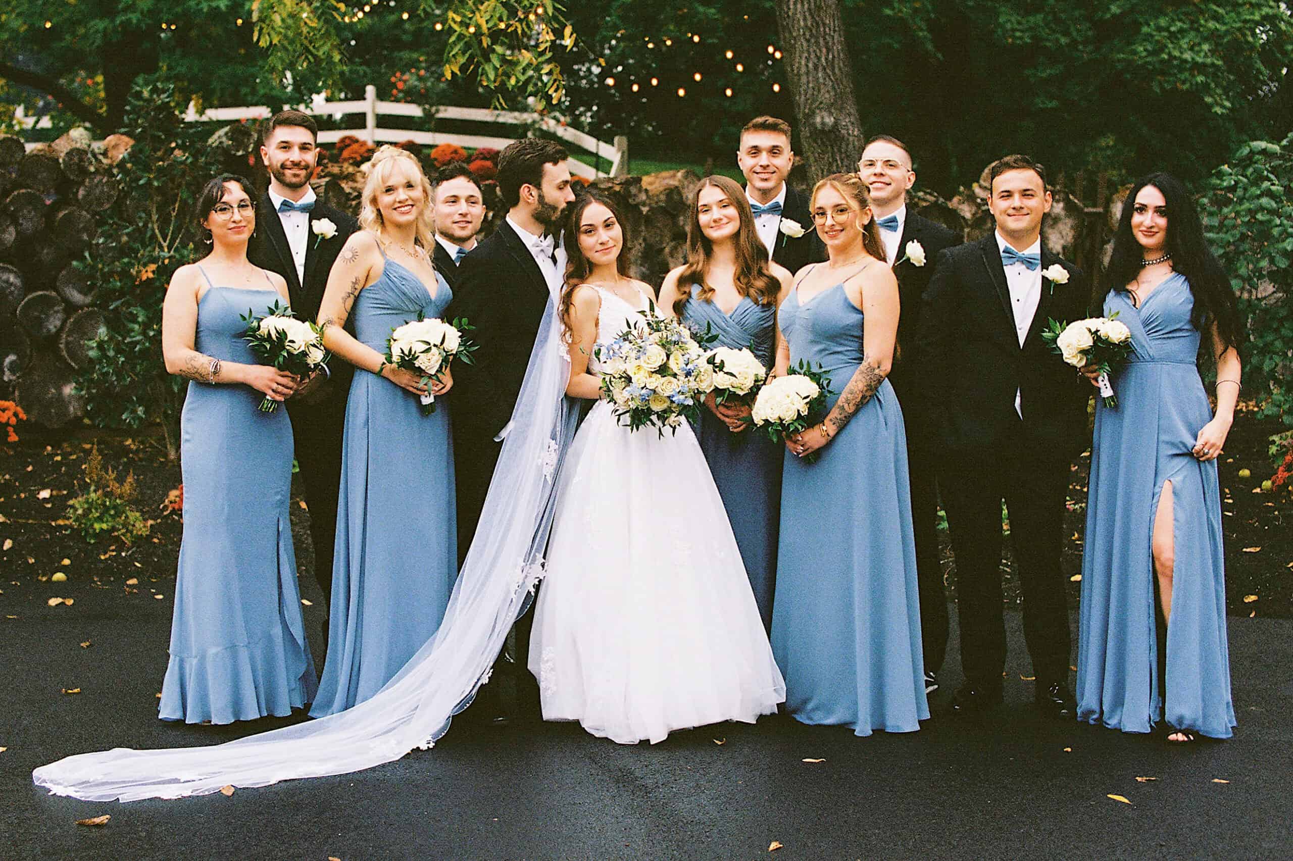 A bride and groom stand with their wedding party outdoors at a beautiful Lochwood Manor wedding. Bridesmaids wear blue dresses, groomsmen wear black suits with bow ties, and everyone holds bouquets or poses for the photo.