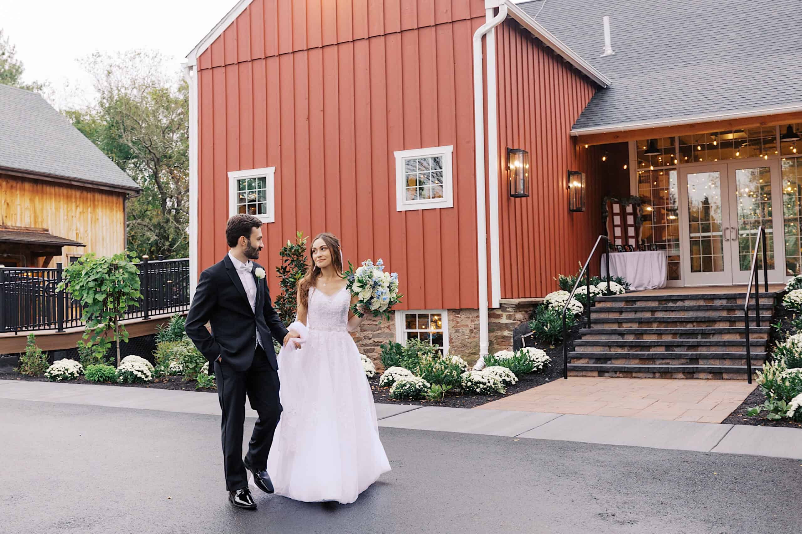 A bride and groom walk outside a red barn-style venue at their beautiful Lochwood Manor wedding, holding hands; the bride wears a white gown and holds a bouquet, while the groom is in a black suit.