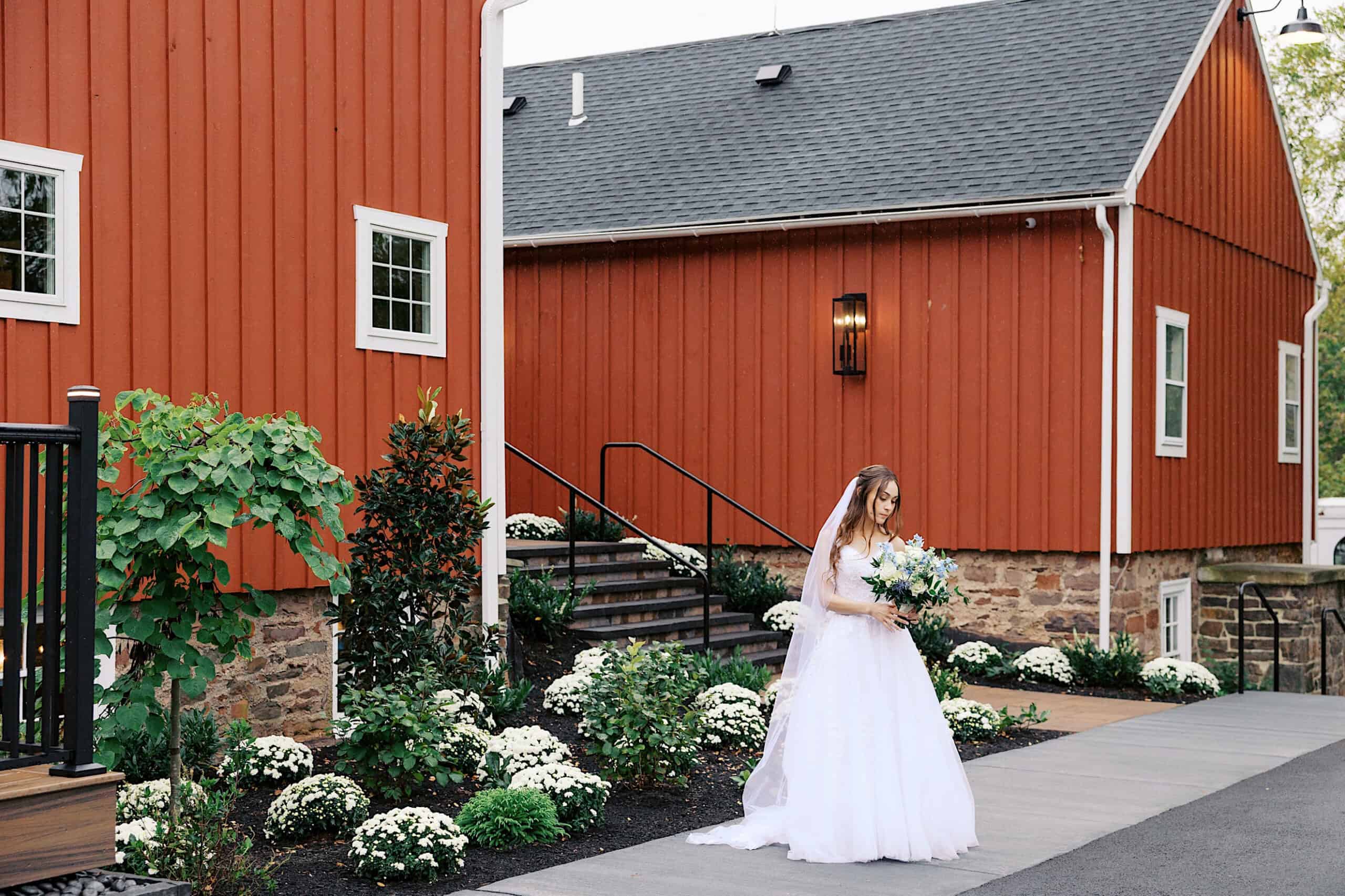 A bride in a white dress holding a bouquet stands outside the beautiful Lochwood Manor, its red building with white trim surrounded by landscaped greenery.