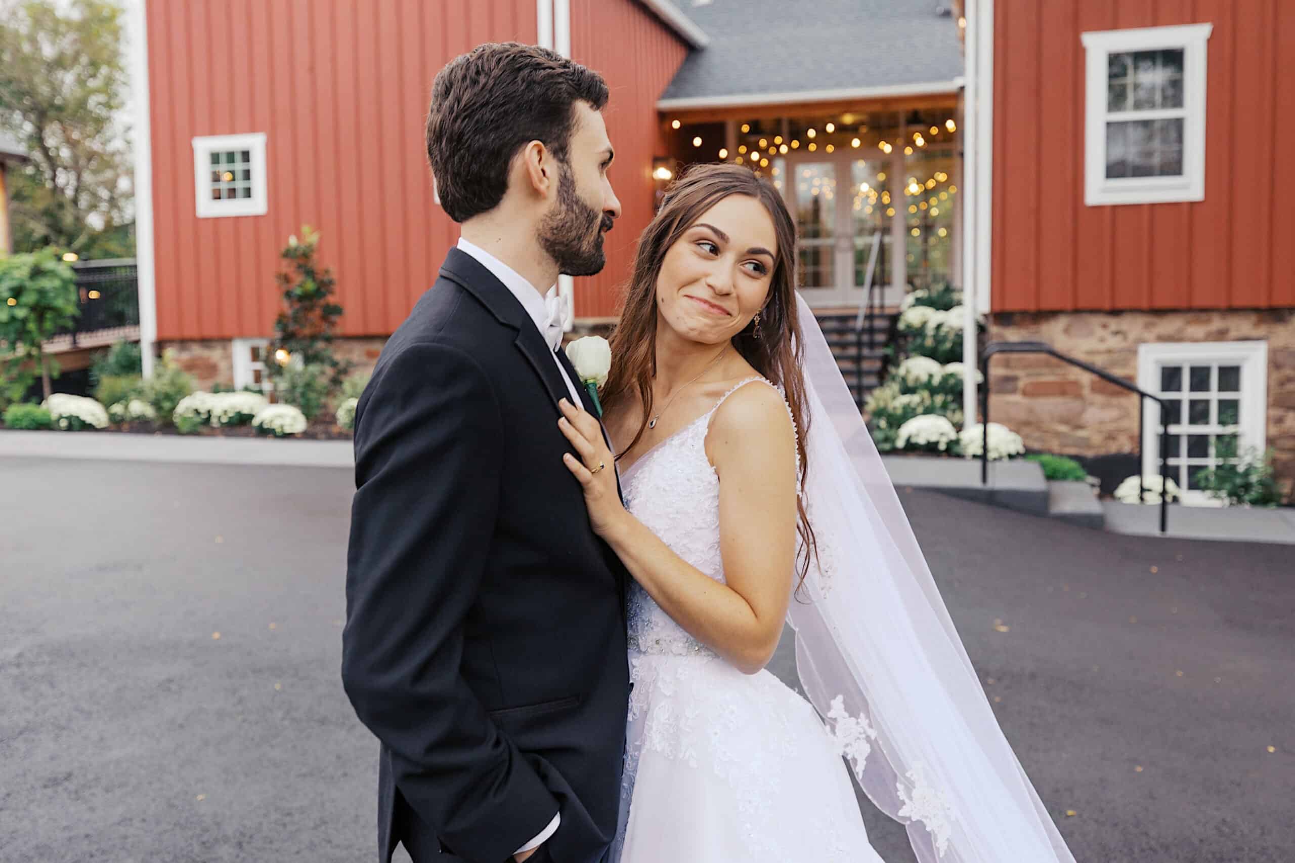A bride and groom stand outside a red barn venue during their beautiful Lochwood Manor wedding; the bride smiles at the groom while holding his lapel, both dressed in wedding attire.