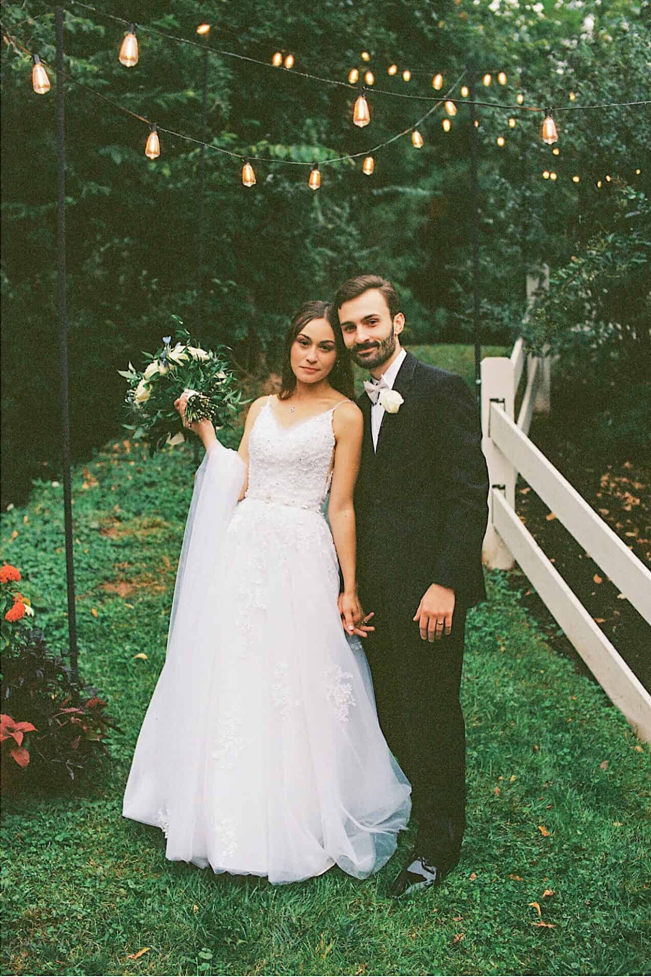 A bride and groom in formal wedding attire walk outdoors near a rustic building and pose together in a garden with string lights overhead during their beautiful Lochwood Manor wedding.