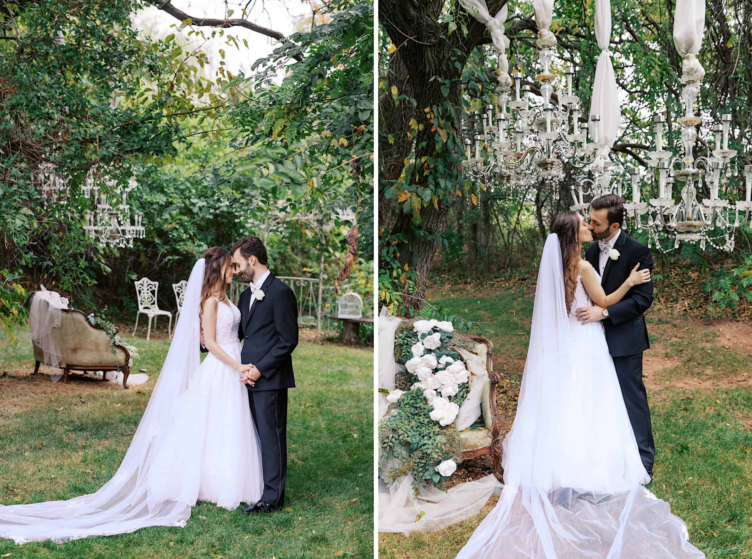 A bride and groom stand together outdoors at their beautiful Lochwood Manor wedding, surrounded by greenery, vintage furniture, and hanging chandeliers, dressed in formal wedding attire.
