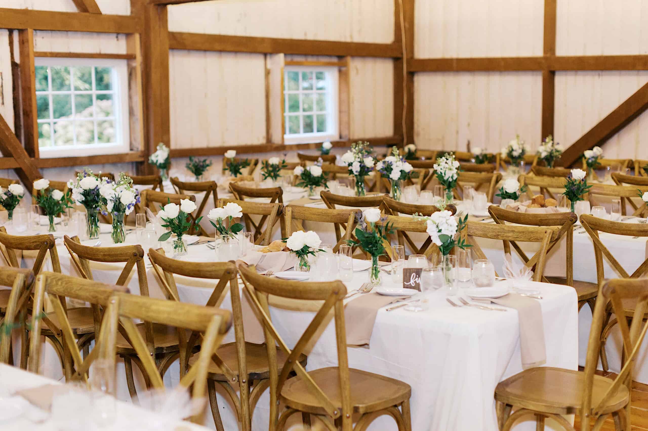 Rows of wooden chairs and rectangular tables set with white tablecloths, glassware, and small white floral arrangements inside a rustic barn-style venue at a beautiful Lochwood Manor wedding.