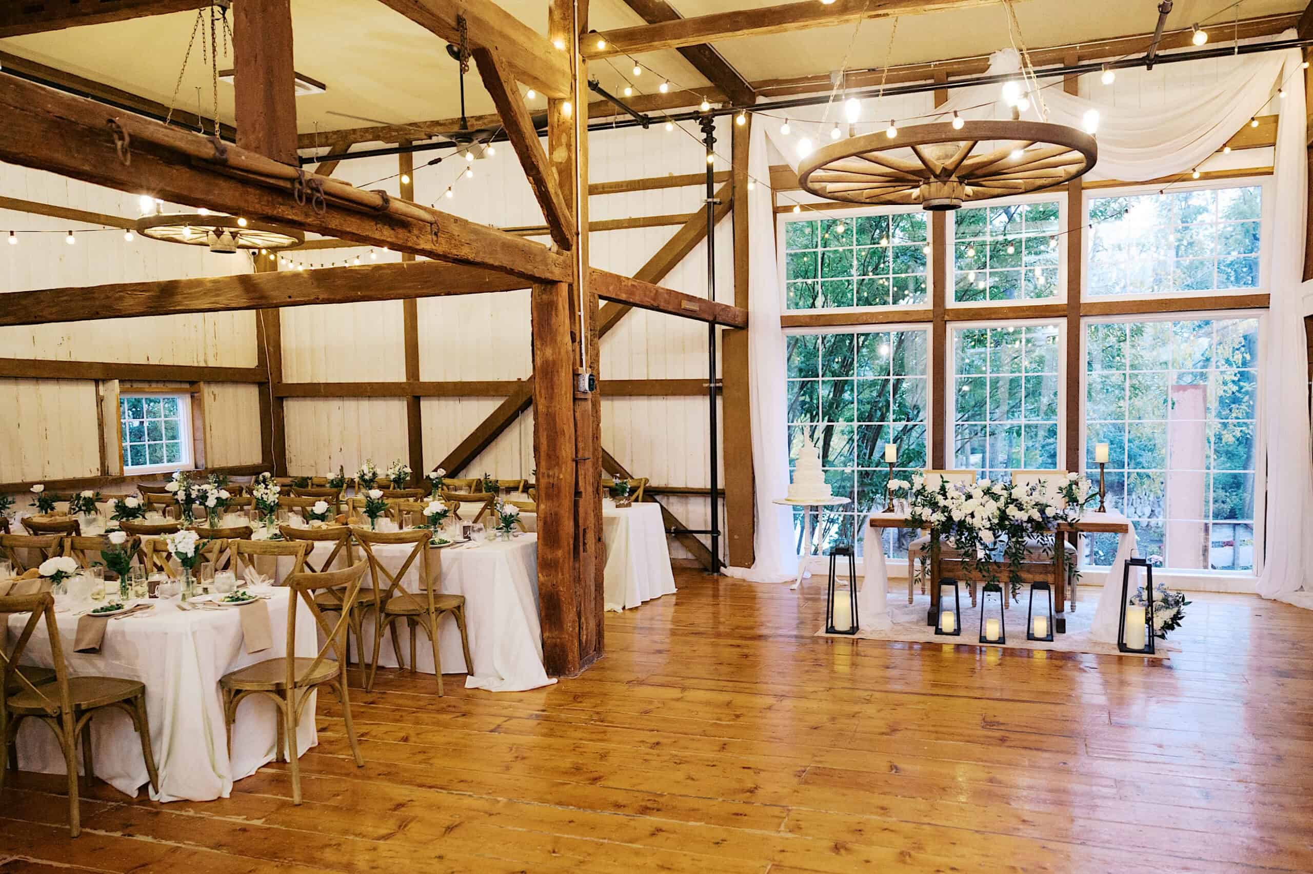 A rustic barn wedding reception setup at beautiful Lochwood Manor features wooden beams, round tables with white tablecloths, floral centerpieces, and large windows letting in natural light.