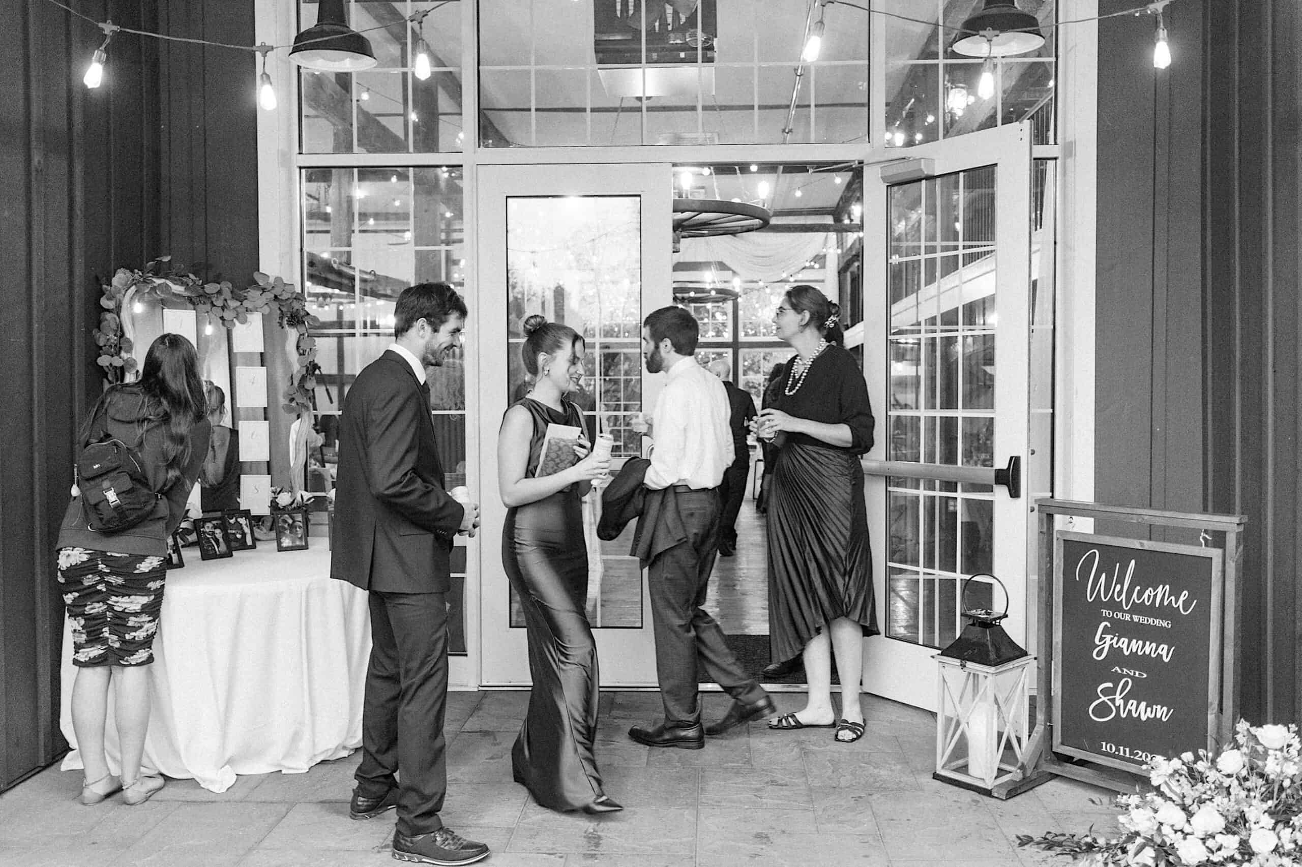 A group of people in formal attire stand and converse at the entrance of a venue with glass doors; a welcome sign and a decorated table hint at a beautiful Lochwood Manor wedding.