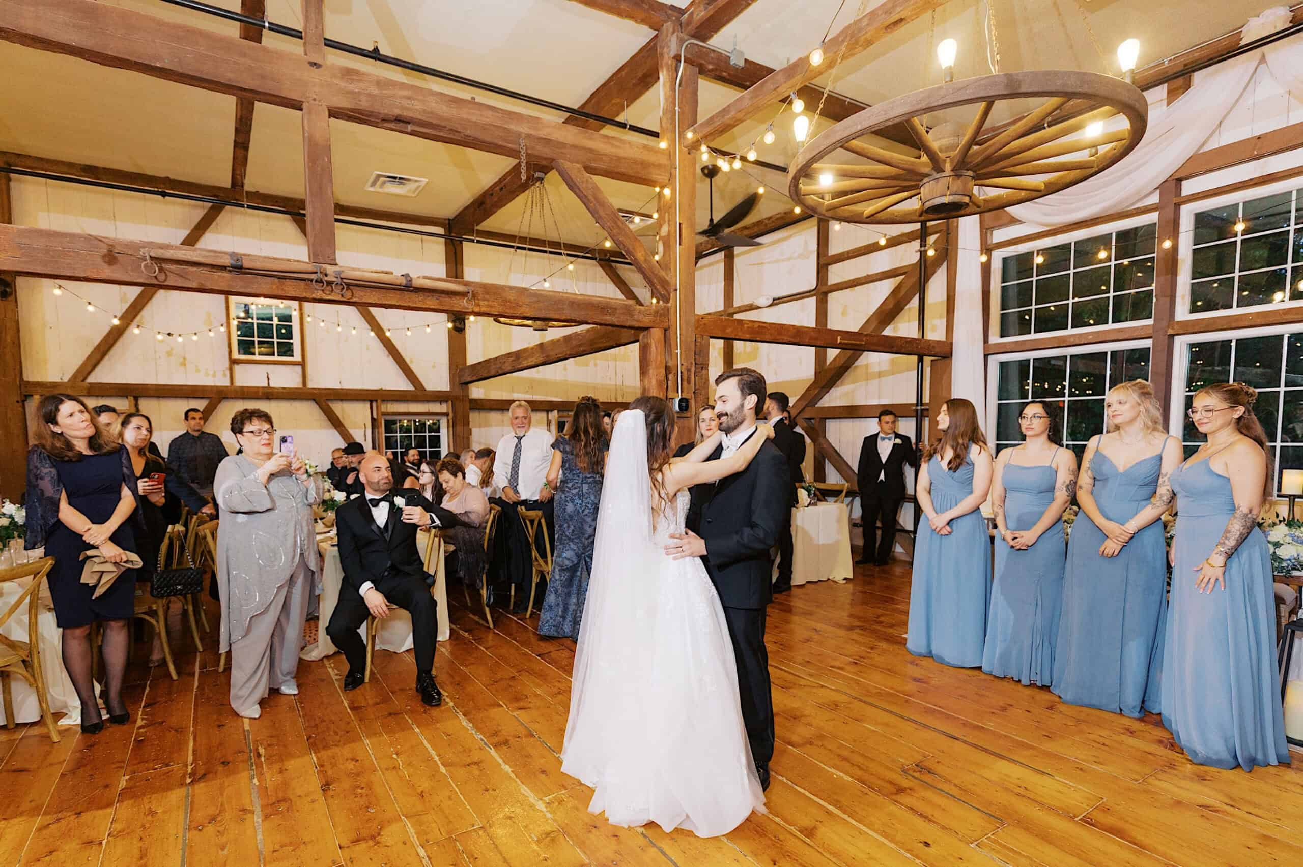 A bride and groom share their first dance in a rustic barn at the beautiful Lochwood Manor wedding venue as guests watch and take photos. Bridesmaids in blue dresses stand to the right.