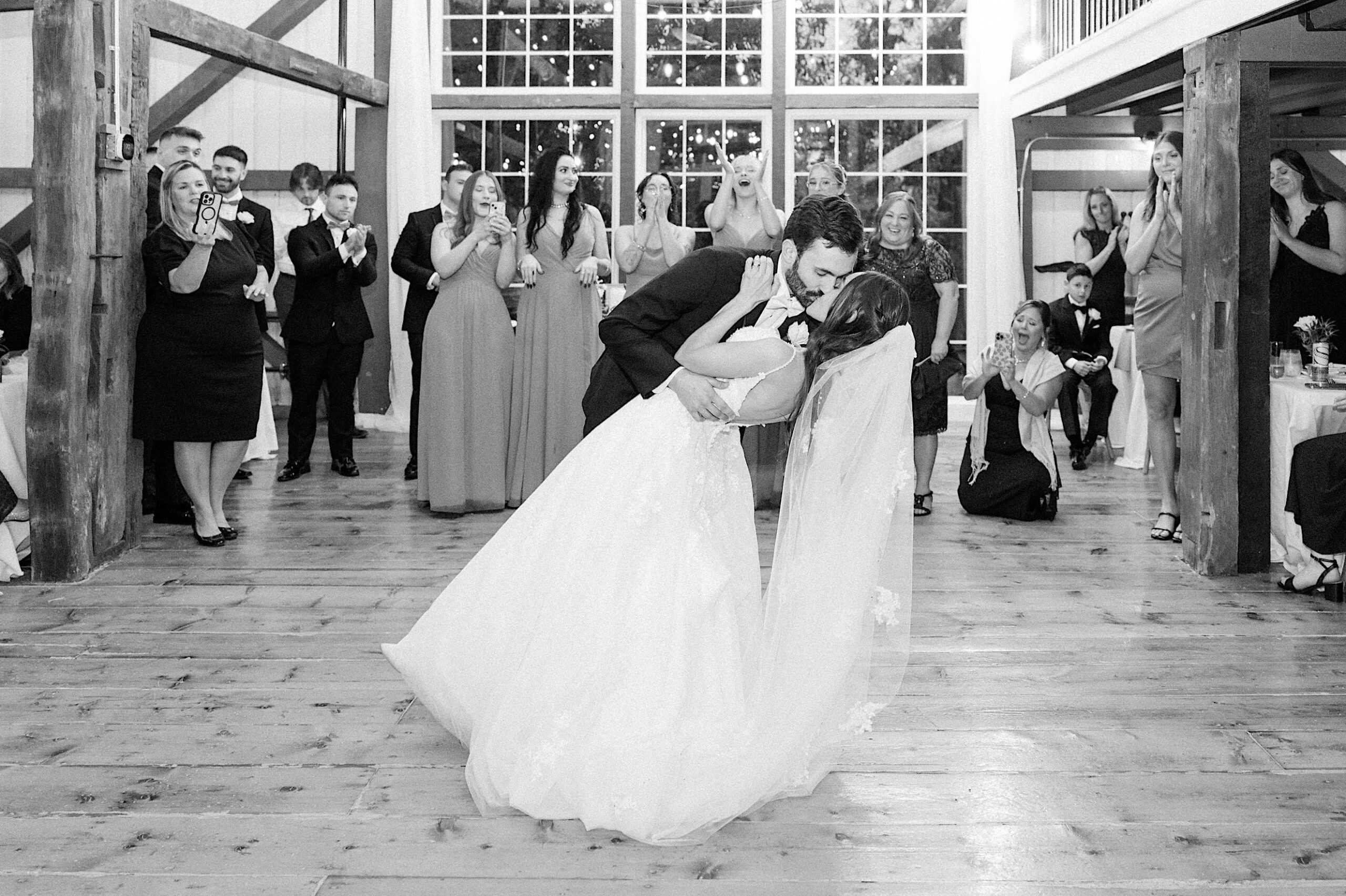 A bride and groom share a dip kiss on the dance floor as guests stand and applaud in a barn-style setting at their beautiful Lochwood Manor wedding.