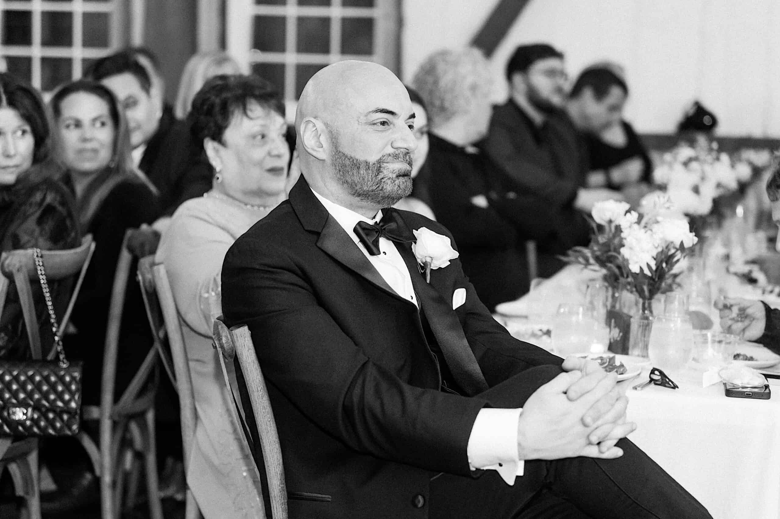 A man in a tuxedo sits with crossed legs at a formal event, surrounded by other seated guests and floral table decorations at a beautiful Lochwood Manor wedding.