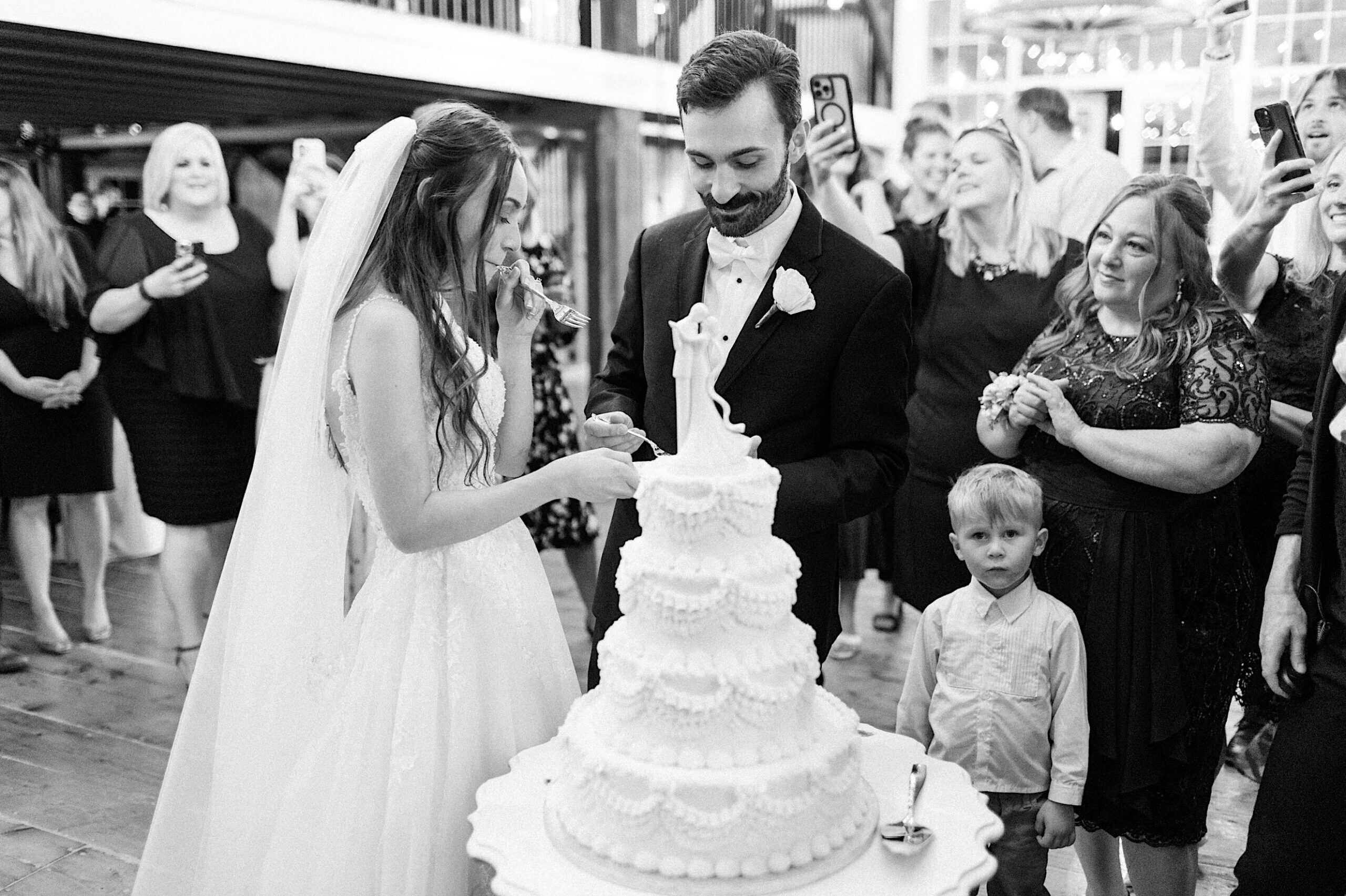A bride and groom cut a tiered wedding cake as guests watch and take photos at a beautiful Lochwood Manor wedding; a small child stands nearby.