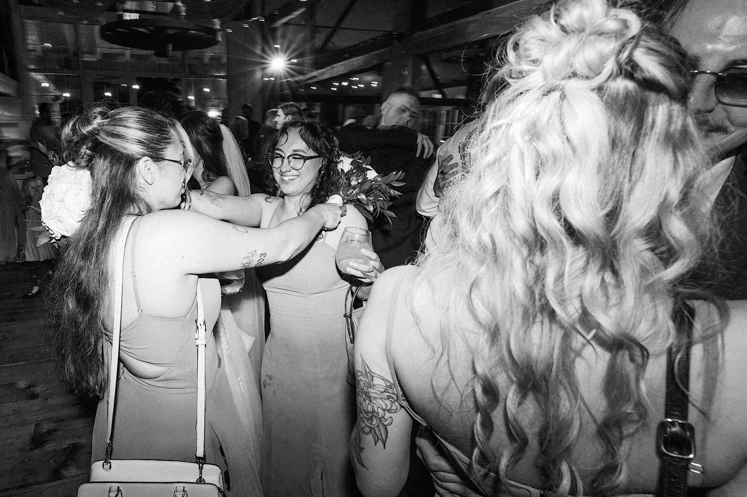 A group of women in dresses interact at a crowded indoor event during a beautiful Lochwood Manor wedding; one holds a bouquet and drink, while others talk and embrace in the background.