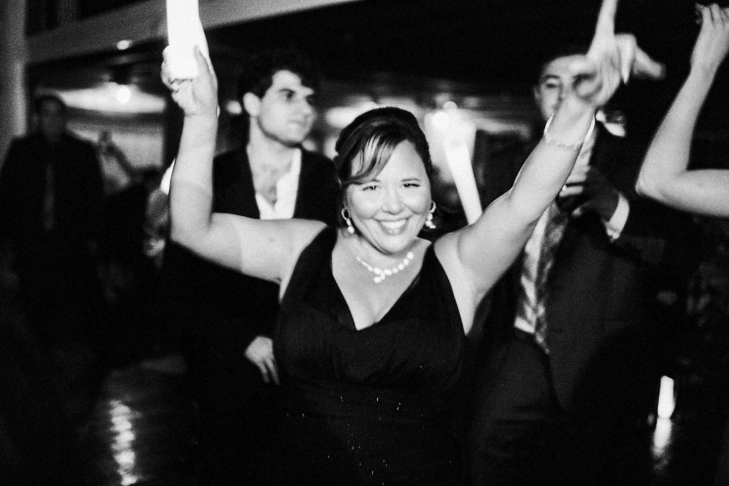 A woman in a black dress smiles with her arms raised while dancing at a beautiful Lochwood Manor wedding, surrounded by other people at the indoor event.