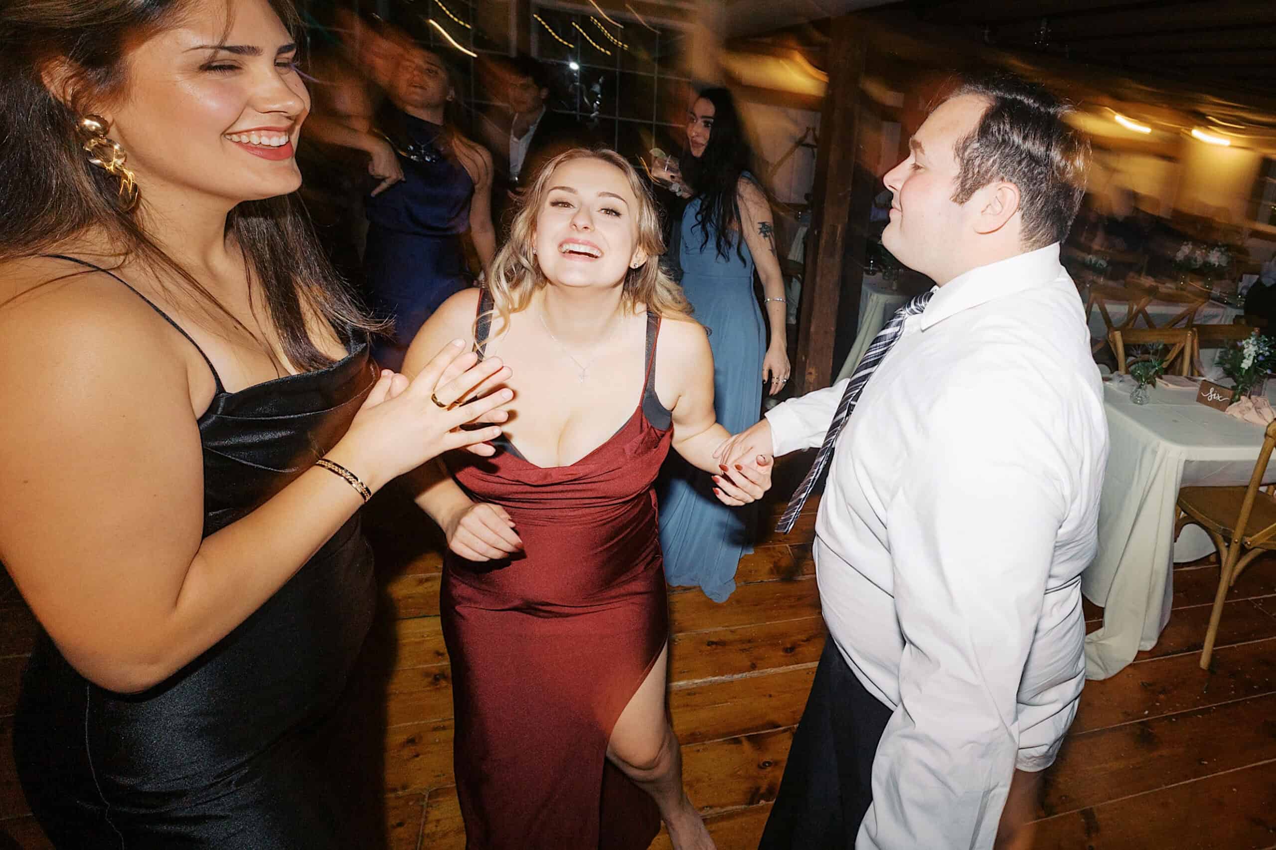 Three adults wearing formal attire smile and dance together on a wooden floor at a beautiful Lochwood Manor wedding, with blurred guests and tables in the background.
