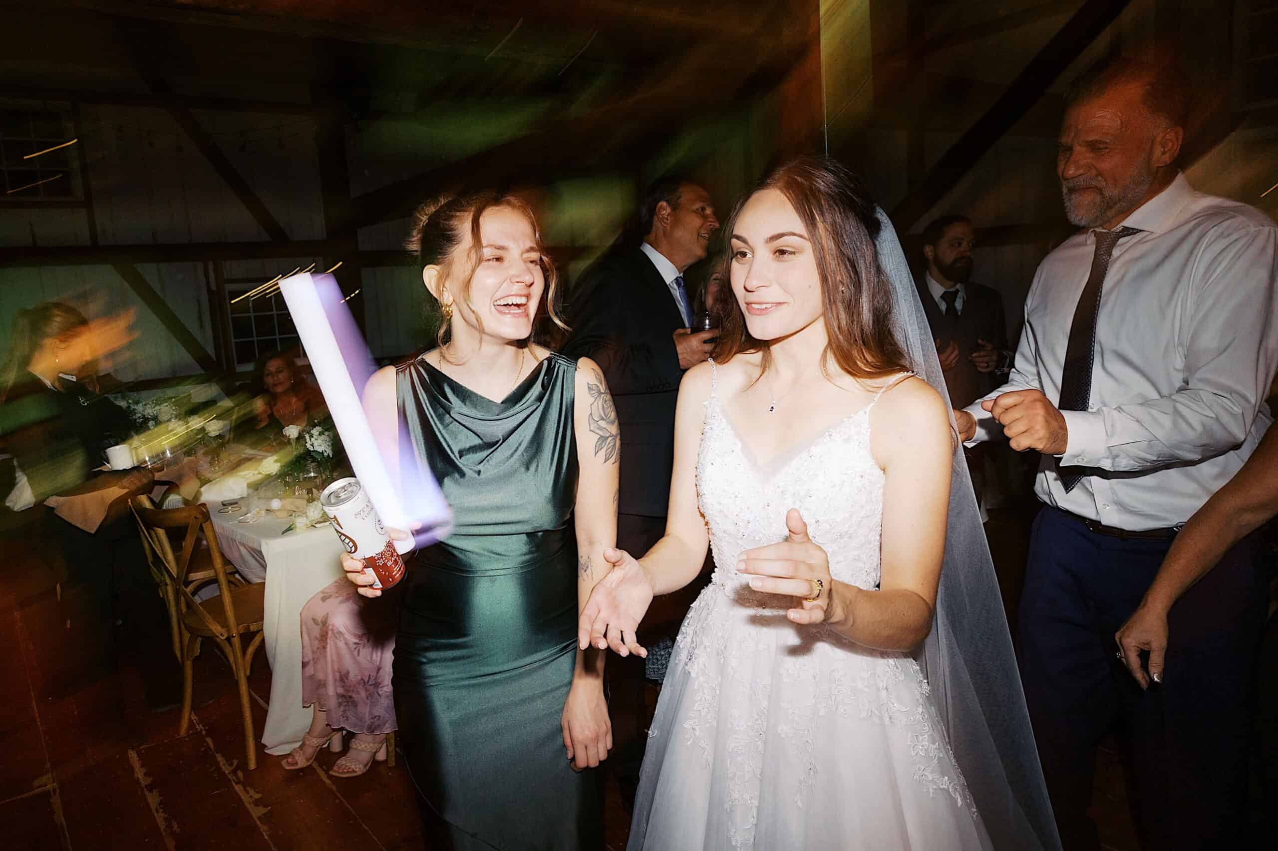 Two women, one in a wedding dress and one in a green dress, stand together smiling on the dance floor at a beautiful Lochwood Manor wedding, surrounded by other guests enjoying the celebration.
