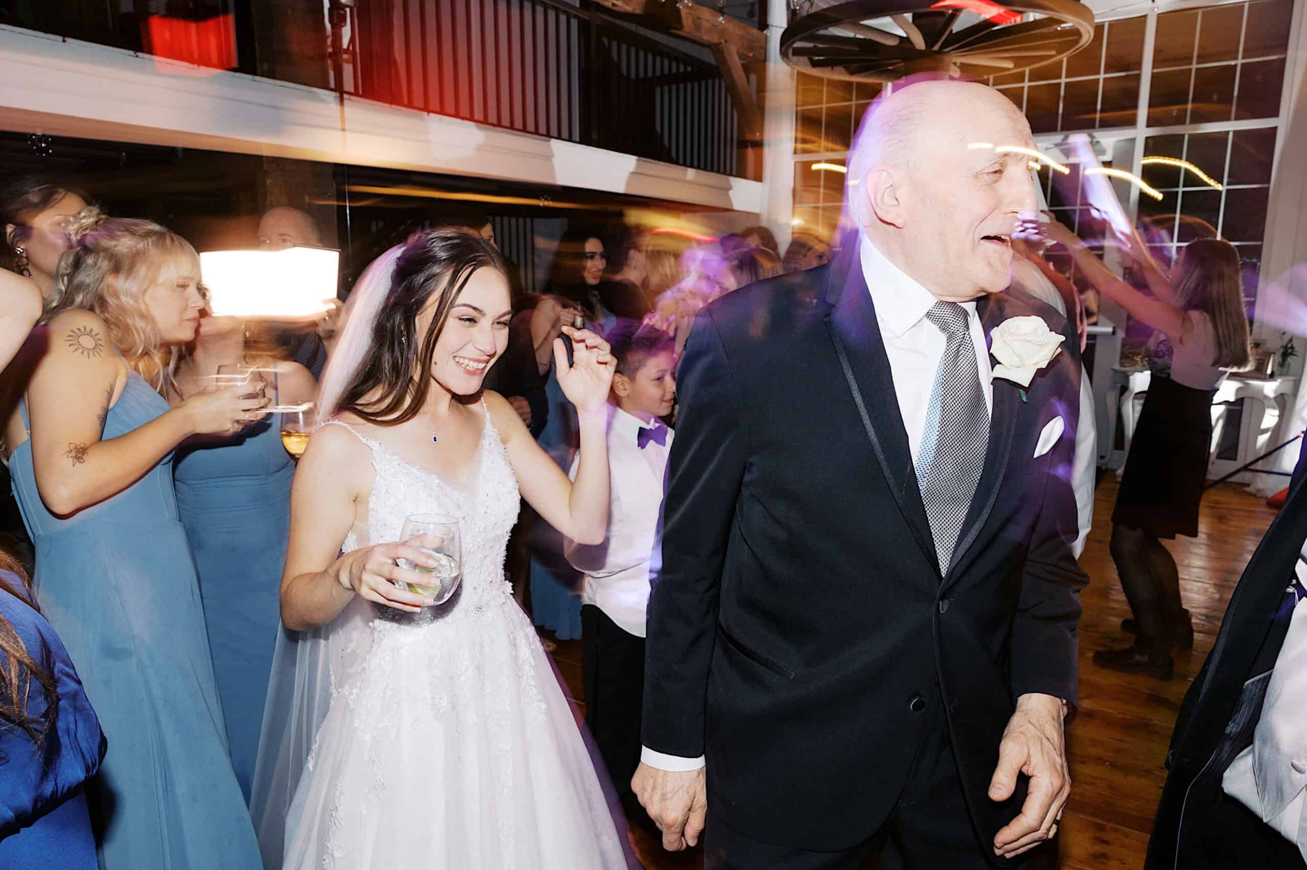 A bride in a white dress and a man in a suit hold hands while dancing at their beautiful Lochwood Manor wedding, surrounded by guests in blue dresses.