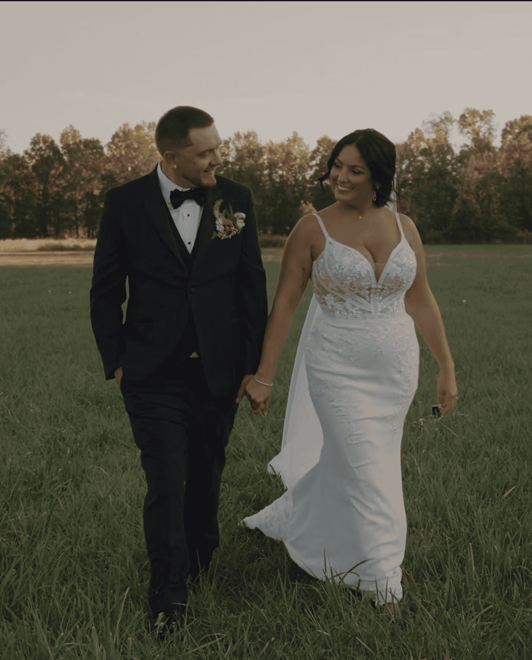 A bride in a white gown and a groom in a black suit walk hand in hand across a grassy field, smiling at each other.