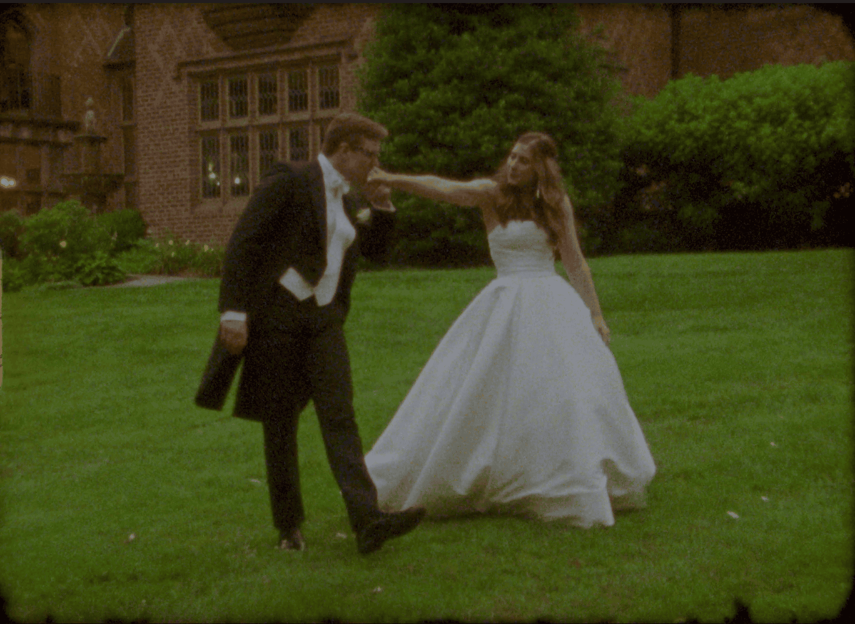 A man in formal attire kisses the hand of a woman in a white wedding dress as they stand on a grassy lawn in front of a brick building.