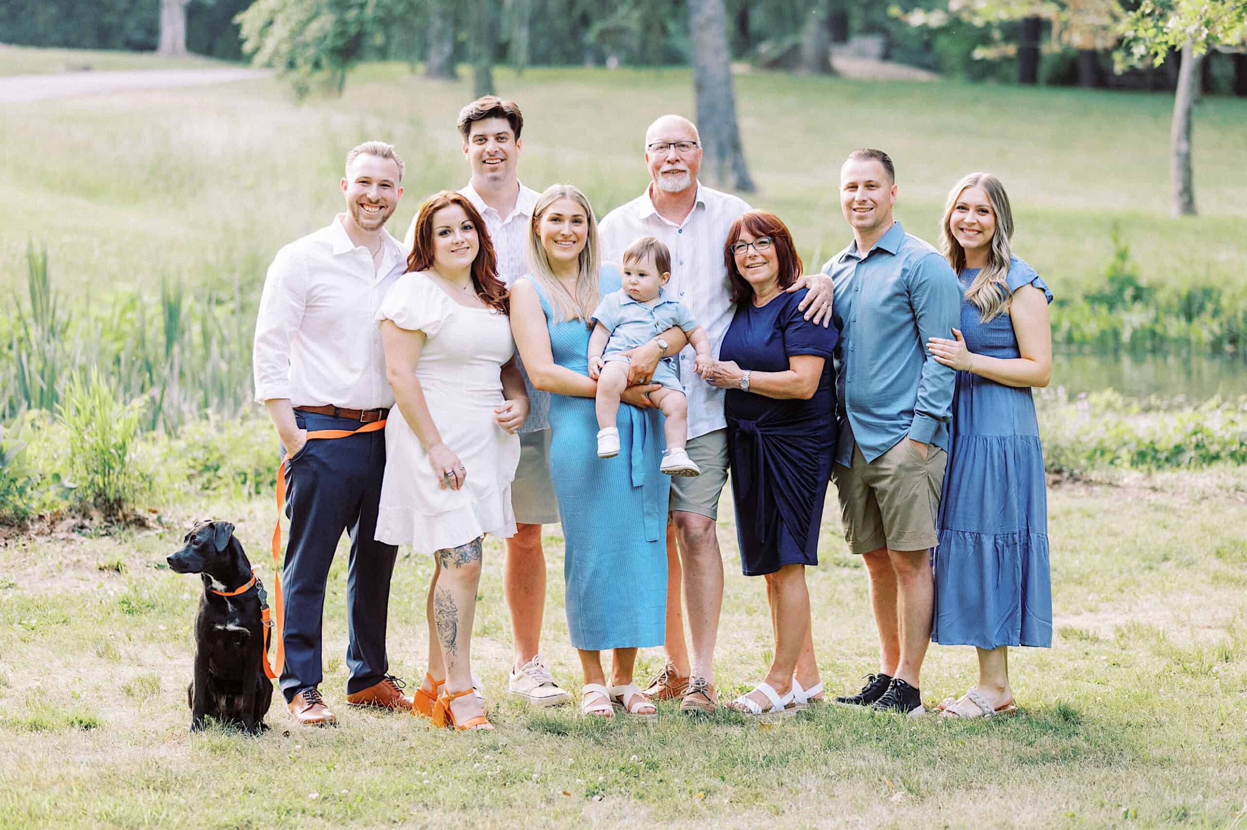 A group of nine adults, one child, and a black dog pose outdoors on grass with trees and a pond in the background, capturing beautiful Spring Family Photos in Bucks County.