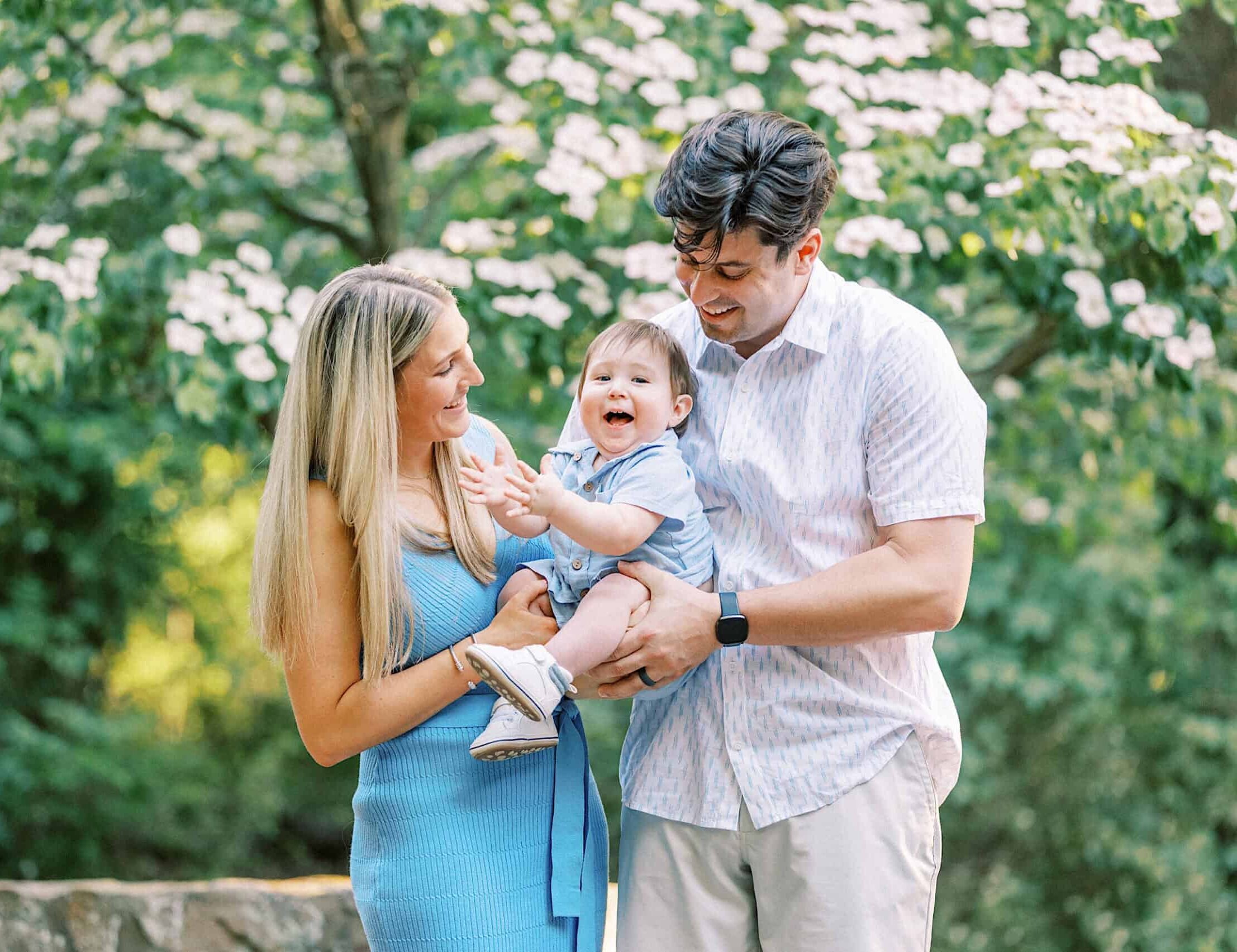 A man and woman stand outdoors, smiling and holding a laughing baby in their arms, surrounded by greenery and flowering trees—capturing joyful Spring Family Photos in Bucks County.