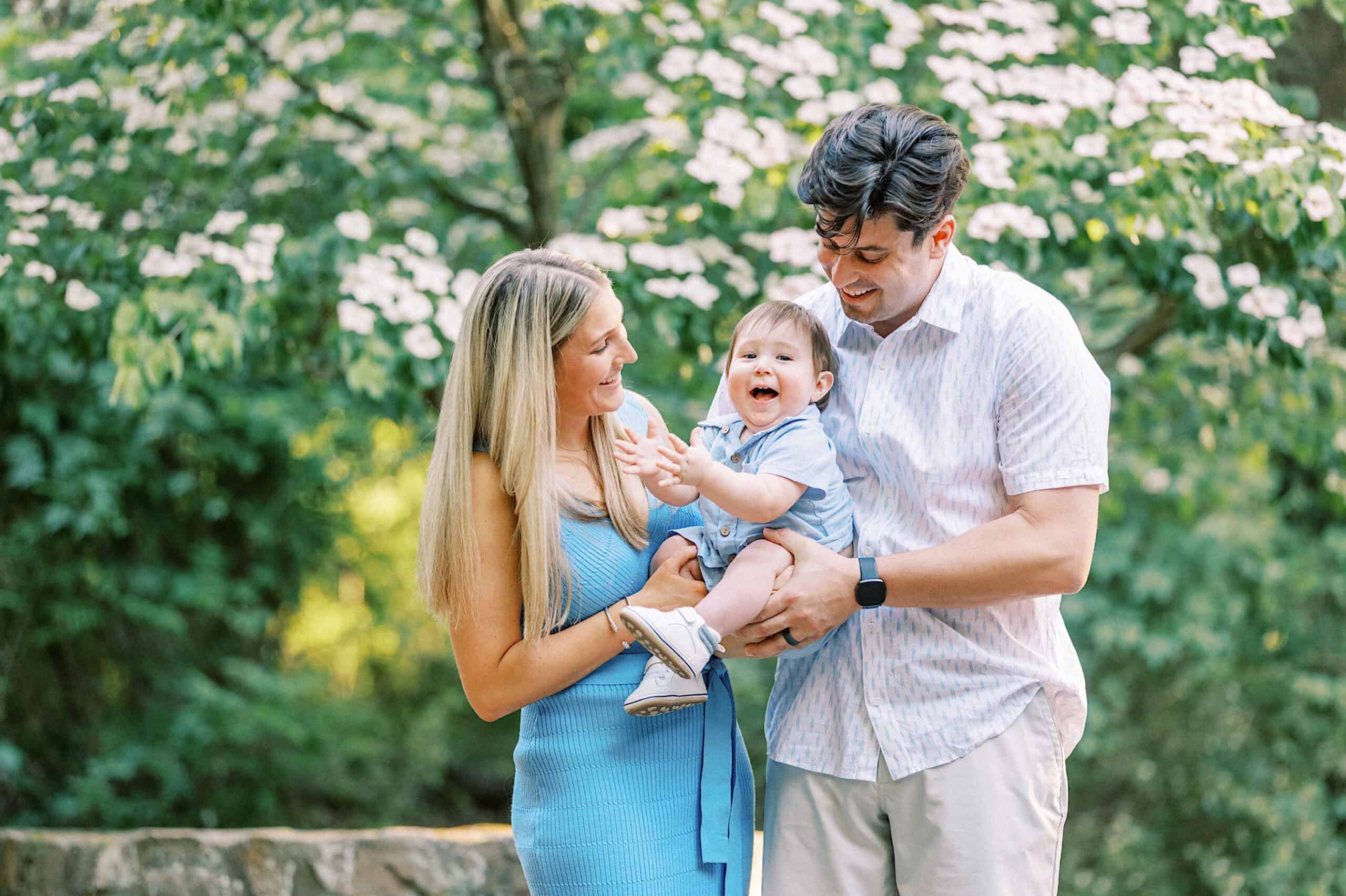 A man and woman stand outdoors, smiling and holding a laughing baby in their arms, surrounded by greenery and flowering trees—capturing joyful Spring Family Photos in Bucks County.