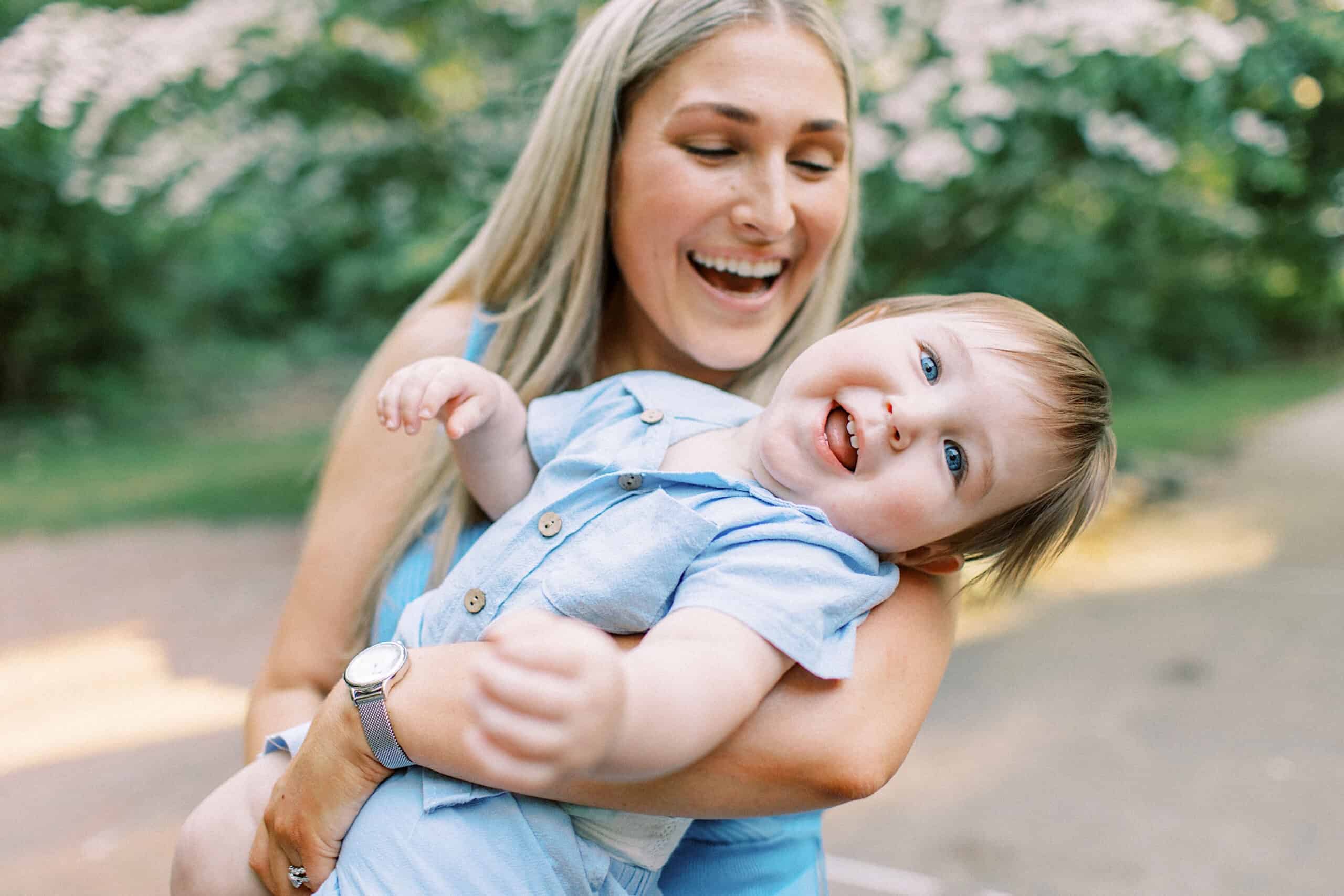 Woman holding a smiling baby outdoors, both in light blue clothing, with greenery in the blurred background—perfect for Spring Family Photos in Bucks County.