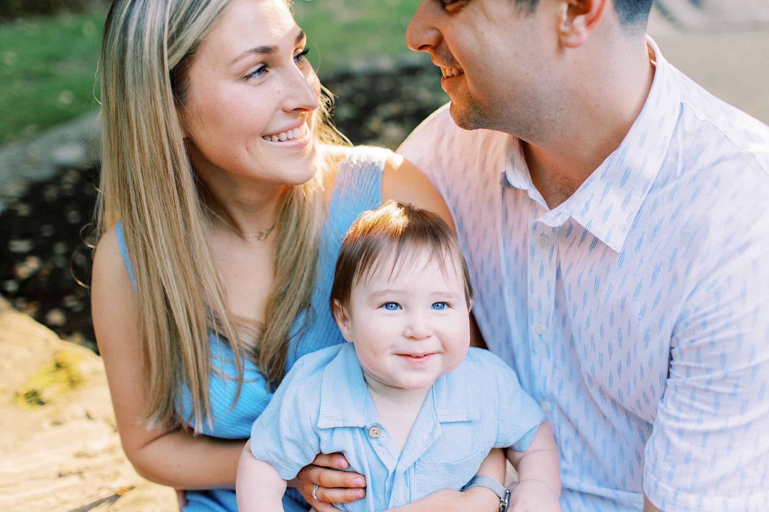 A woman and man sit close together, smiling at each other, while a baby in a blue outfit sits in the woman's lap, looking at the camera during Spring Family Photos in Bucks County.
