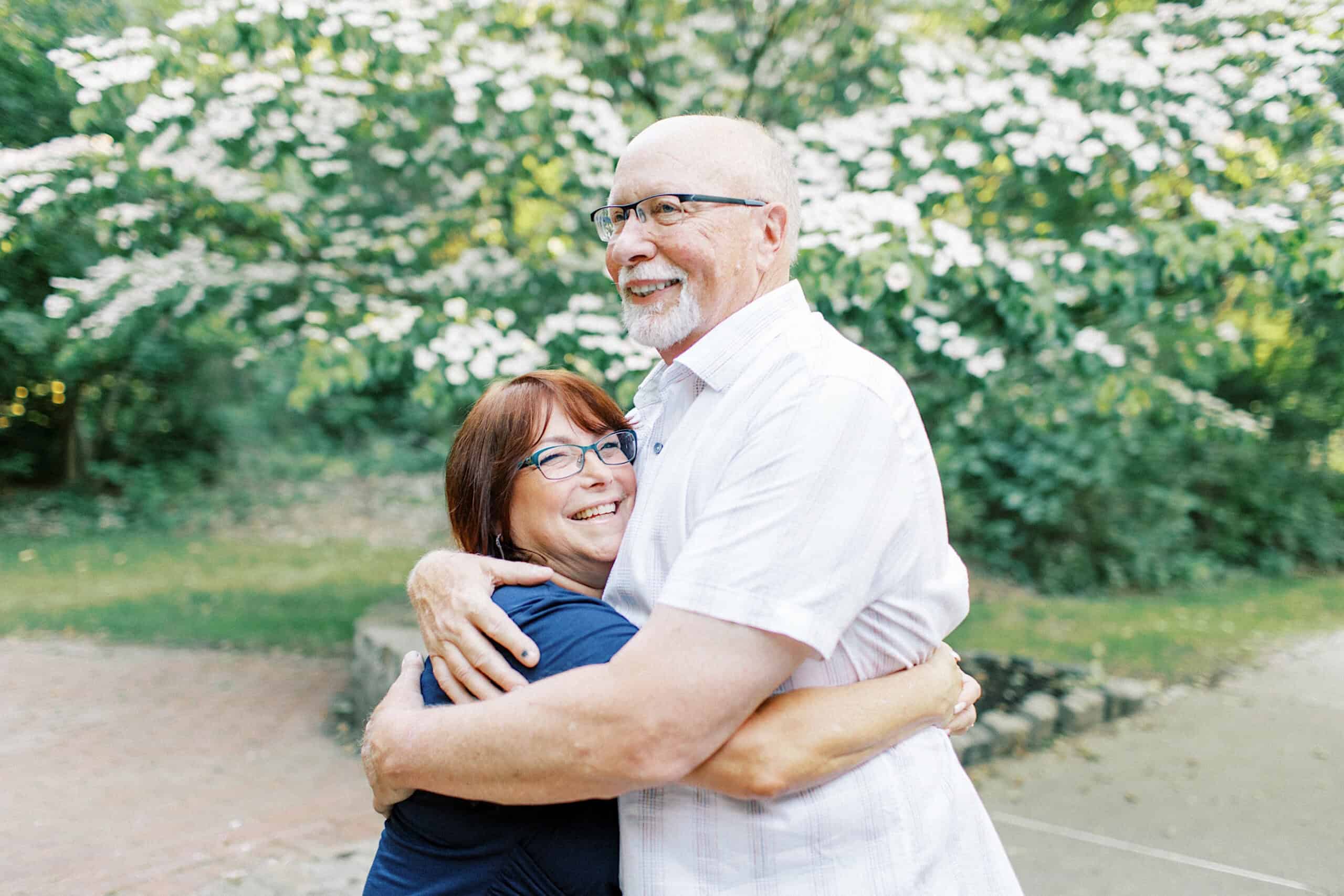An older man and woman stand outdoors, smiling and hugging each other among trees and blooming flowers—capturing the warmth of Spring Family Photos in Bucks County.